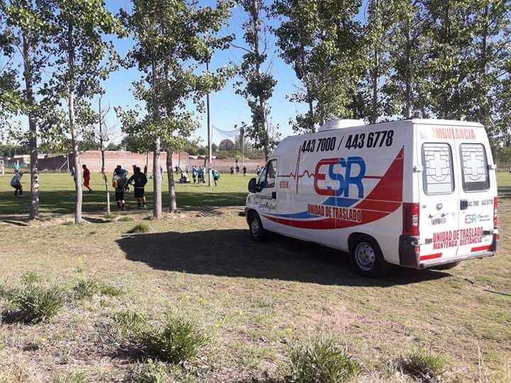 Una ambulancia estacionada sobre el césped junto a un campo donde la gente practica deporte en un día soleado.