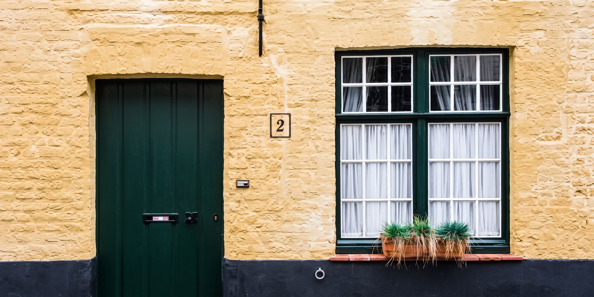 Yellow stucco house facade with a dark green door and a window with white curtains and flower box