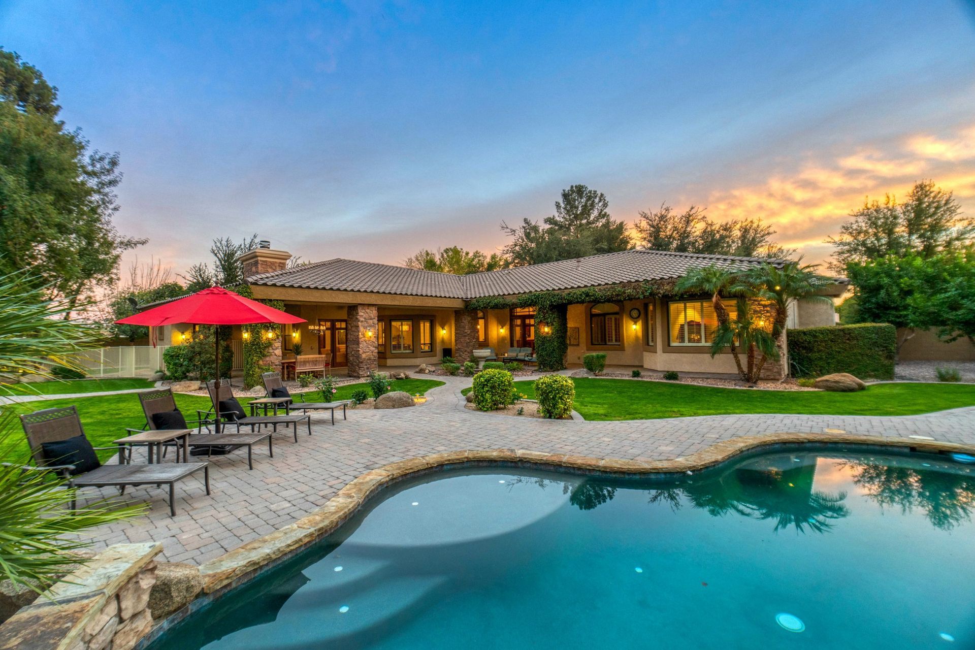 Backyard pool beside a stone patio, lawn chairs, red umbrella, and a lit house at sunset