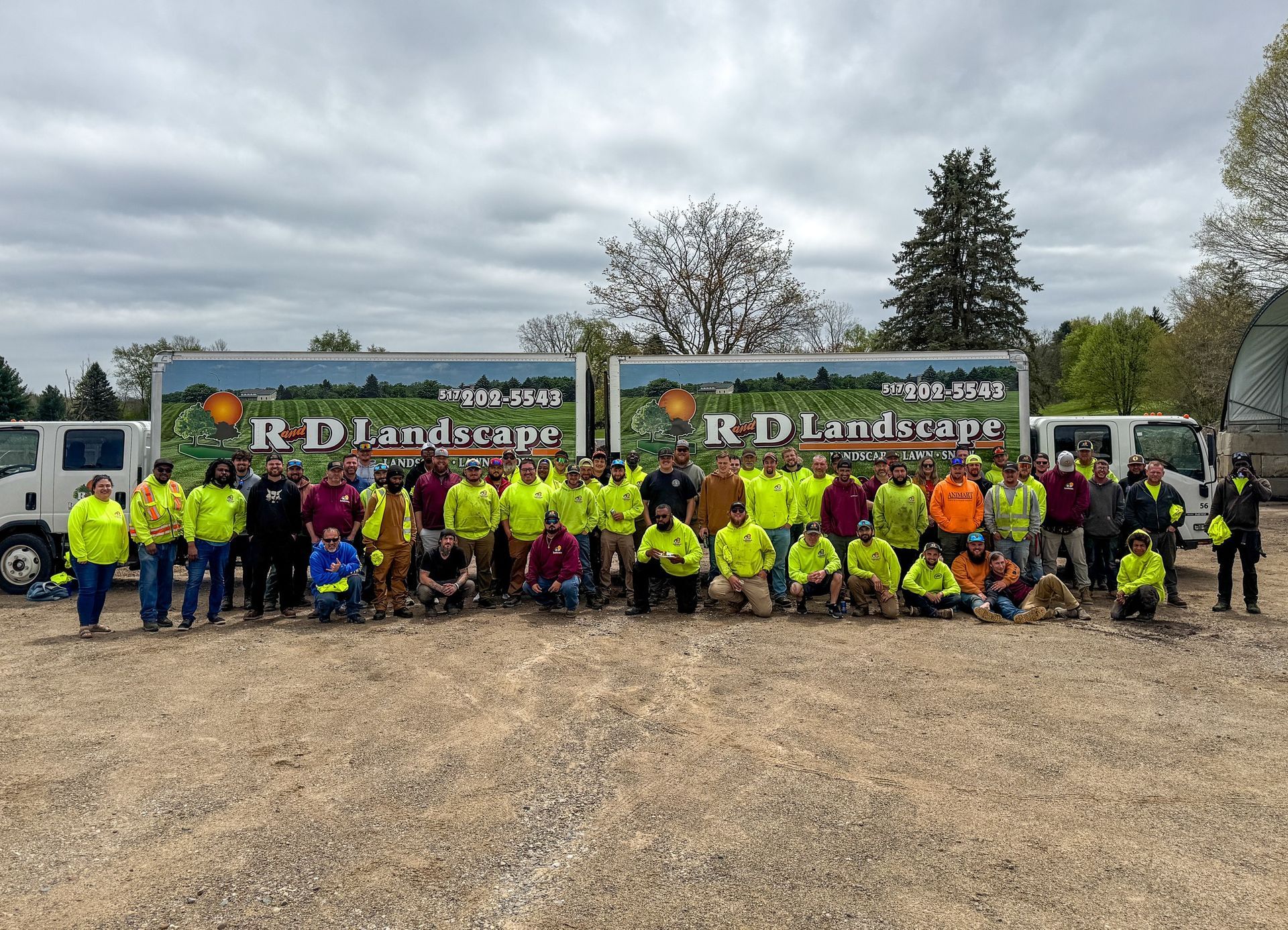 Large group in hi-vis jackets posing in front of two R D Landscaping trucks on a gravel lot under cloudy skies