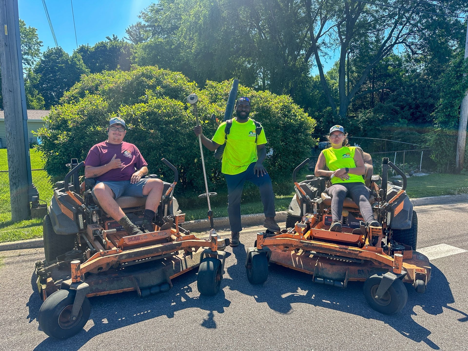 Three workers on large ride-on mowers in a sunny park, wearing neon safety vests.