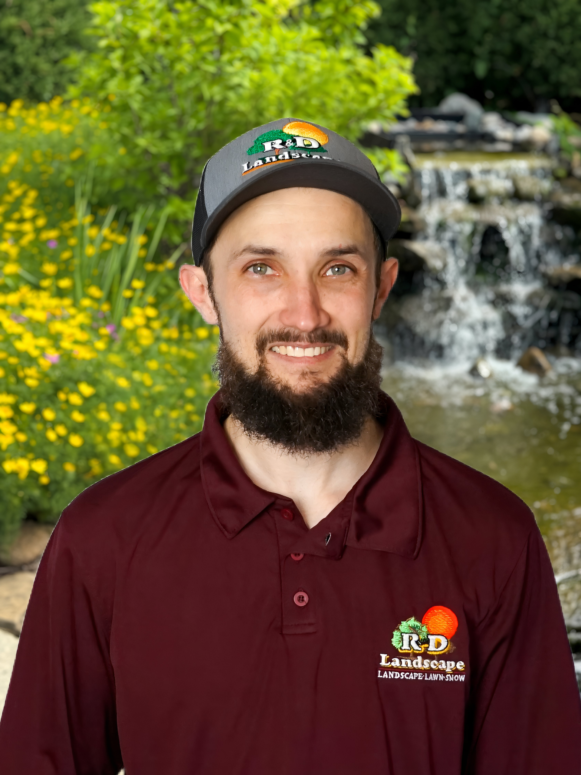 Man in maroon polo and cap smiling in front of a waterfall and yellow flowers