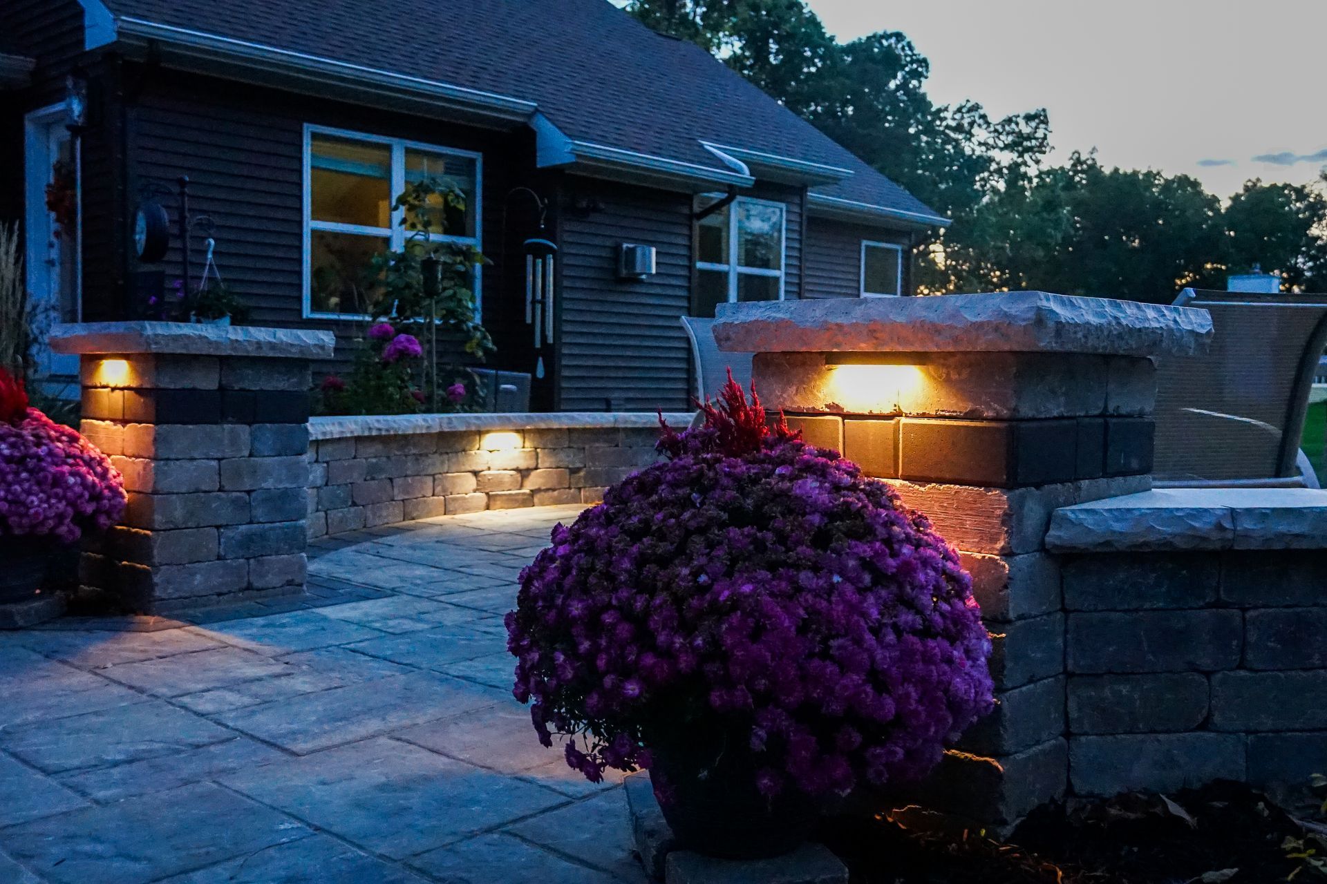 Modern home exterior at dusk with glowing stone walls, paved patio, and purple shrubs