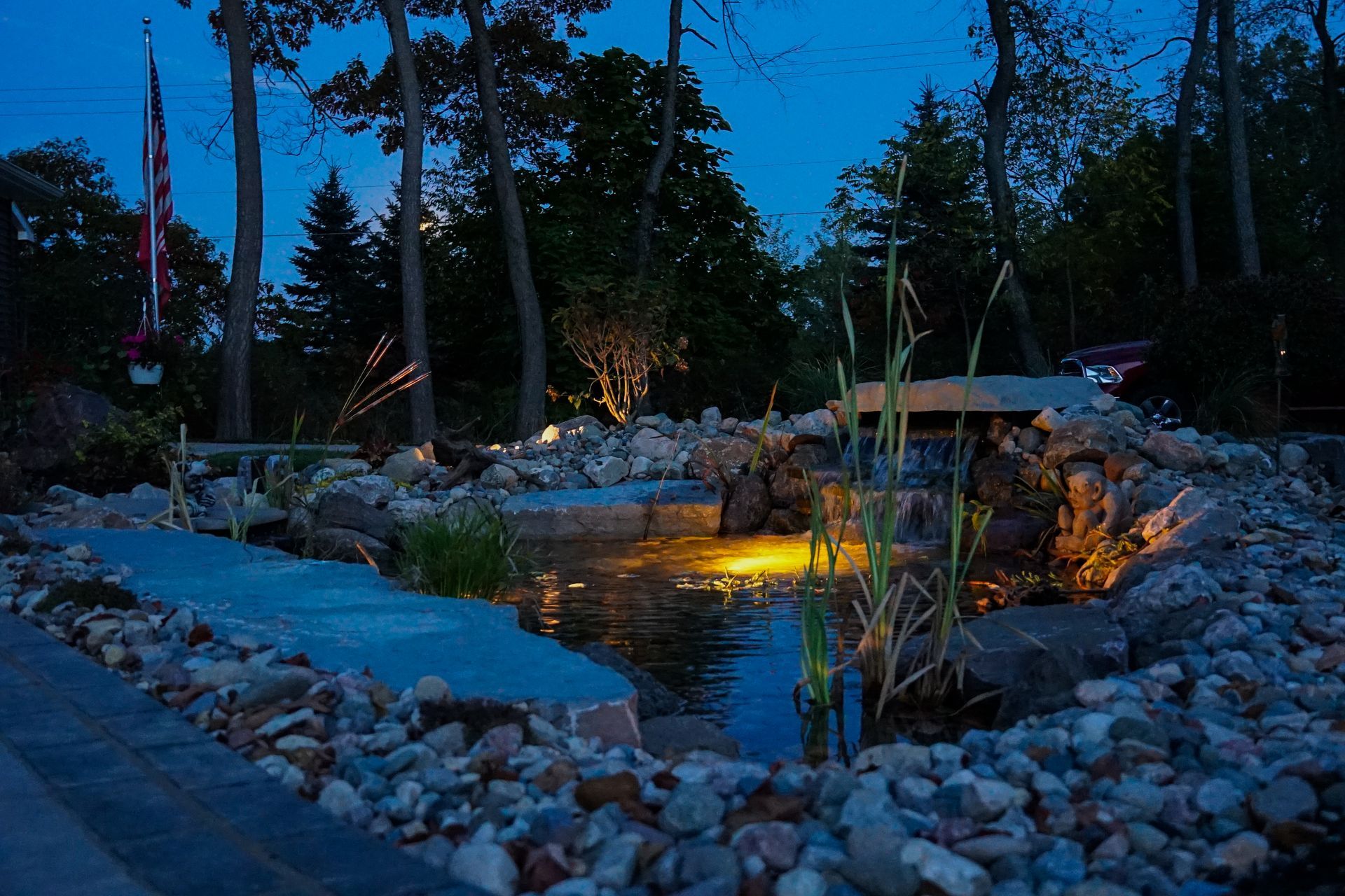 Stone-lined backyard pond at dusk with glowing lights and tall trees nearby