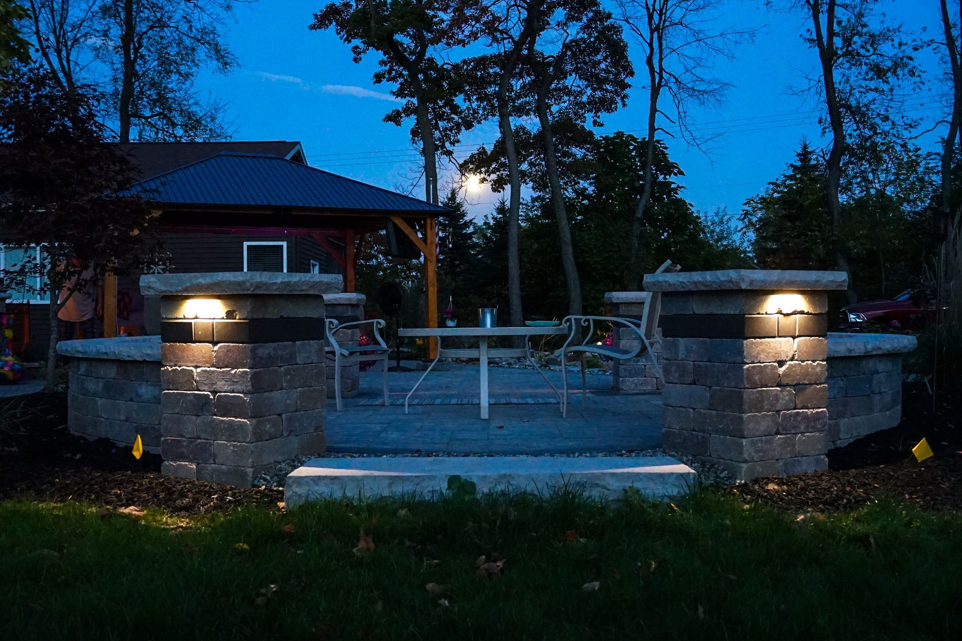 Stone patio entrance at dusk with lit pillars, steps, and trees in the background