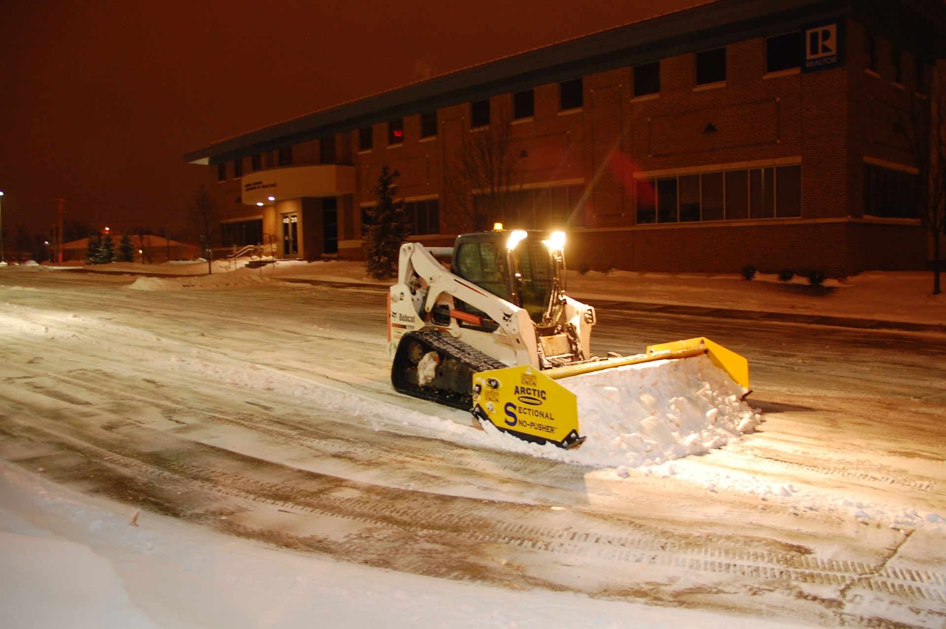 Snowplow clearing a snowy street at night beside a building, with bright headlights and orange-lit sky