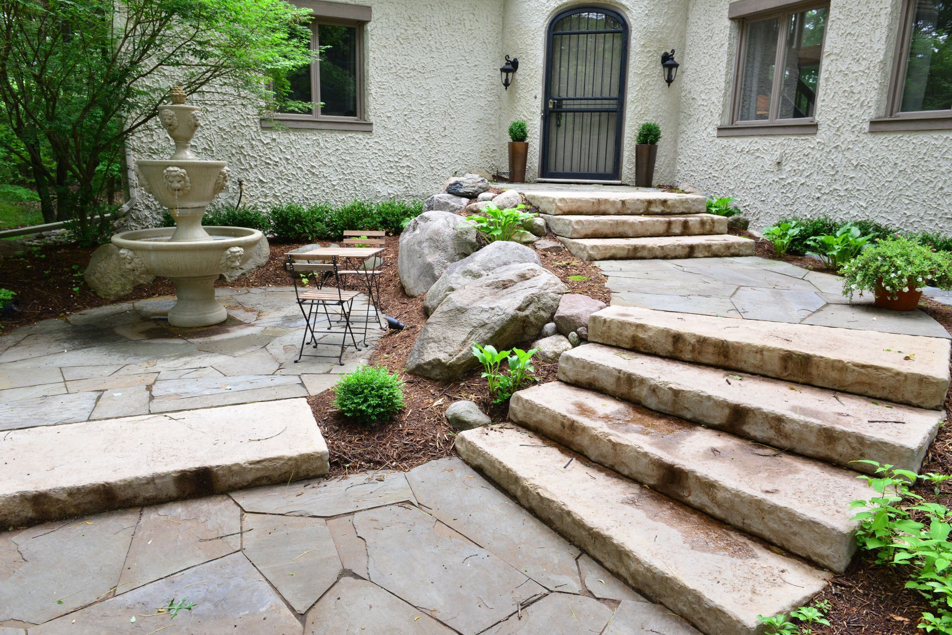 A stone patio with stairs leading up to a fountain in front of a house.