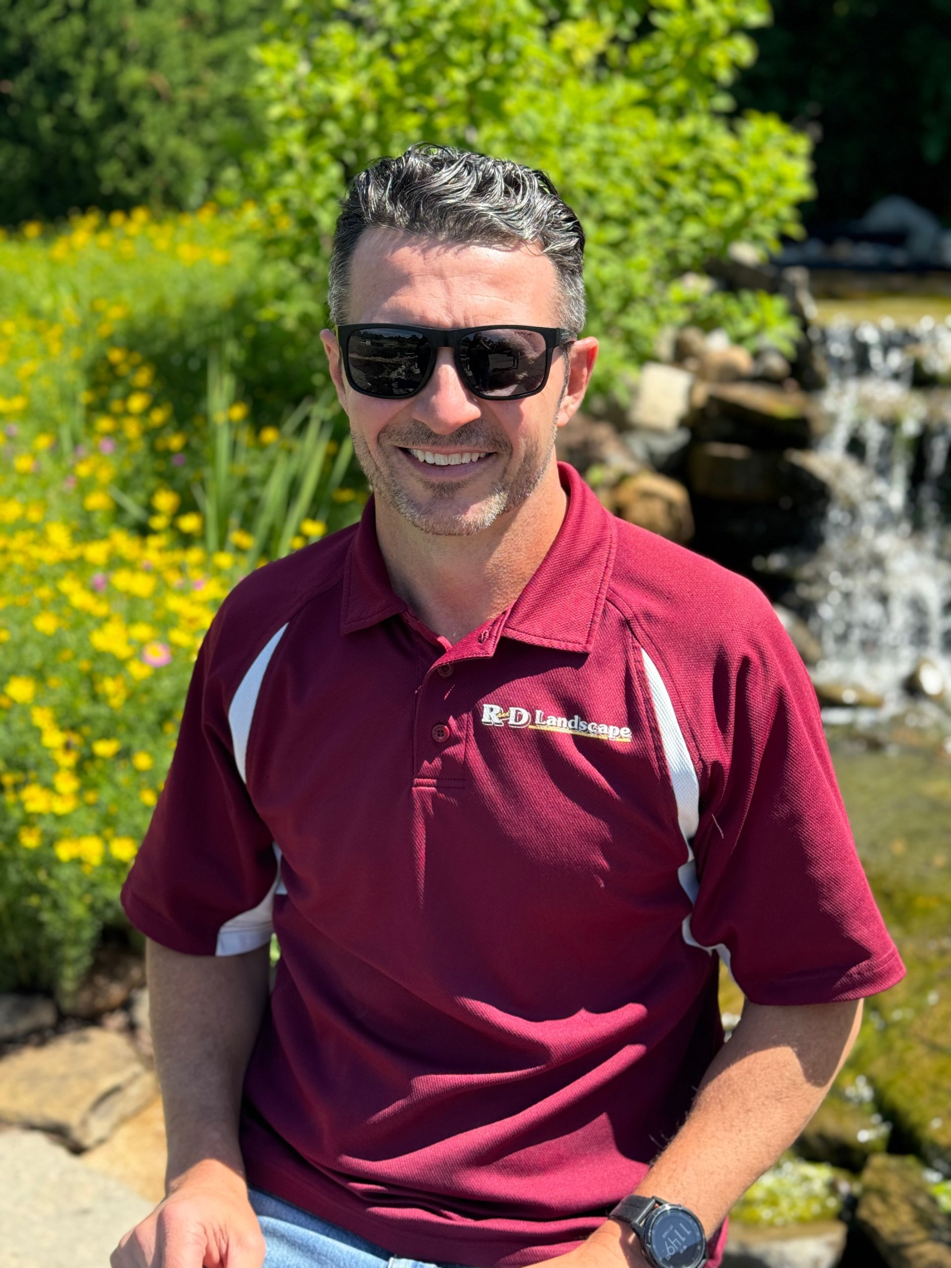 A man wearing sunglasses and a maroon polo shirt is sitting in front of a waterfall.