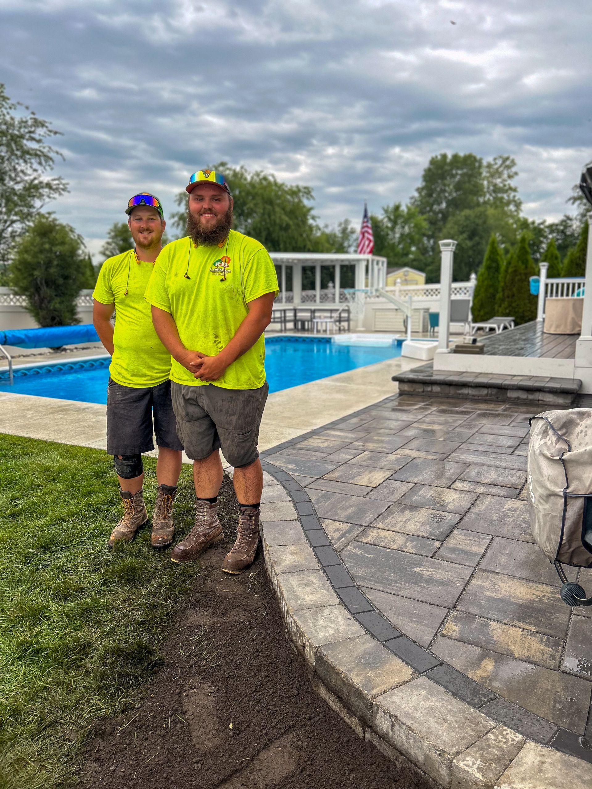 Two people in neon yellow shirts standing beside a pool and stone patio under a cloudy sky