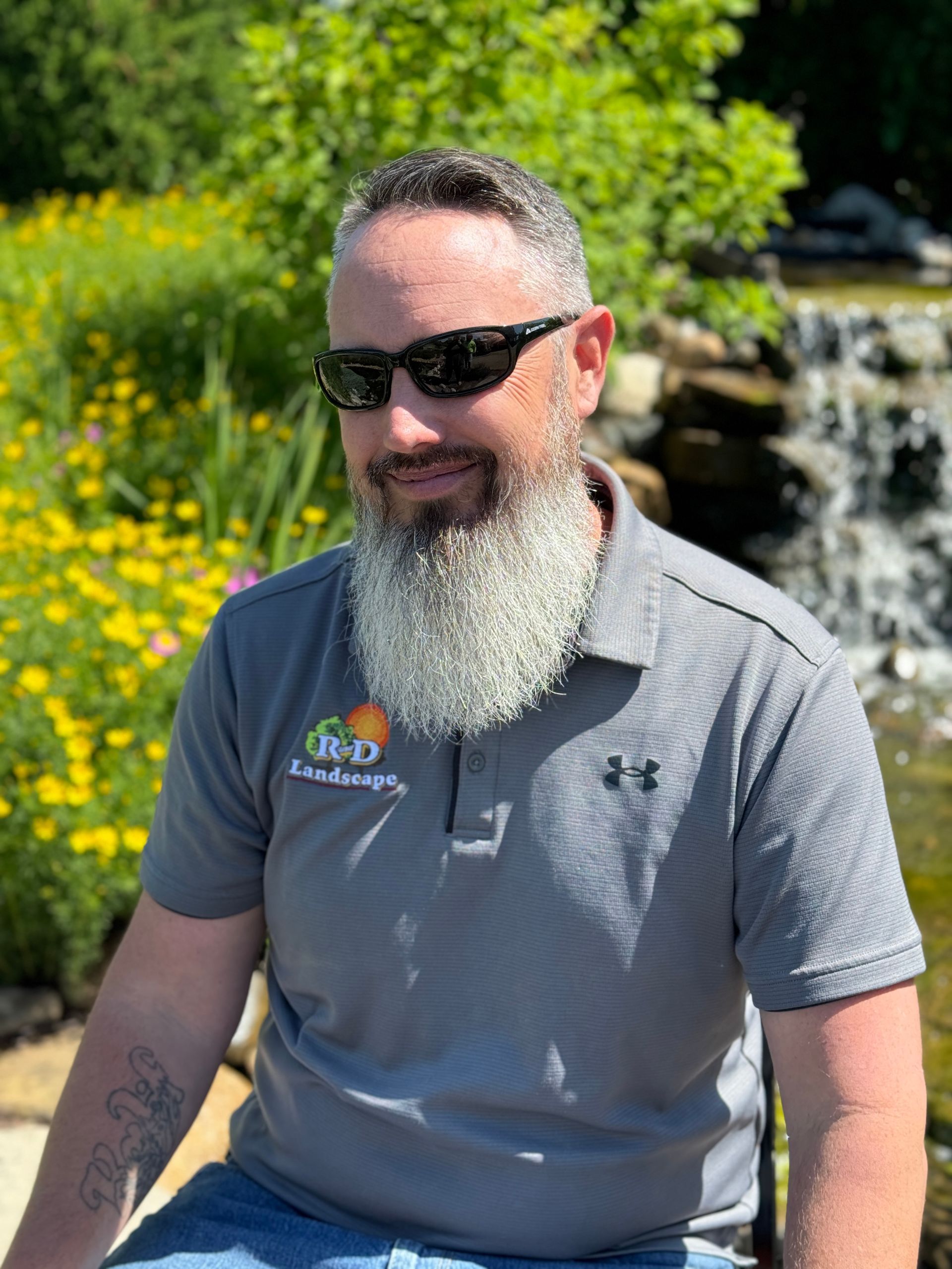 A man with a beard and sunglasses is sitting in front of a waterfall.