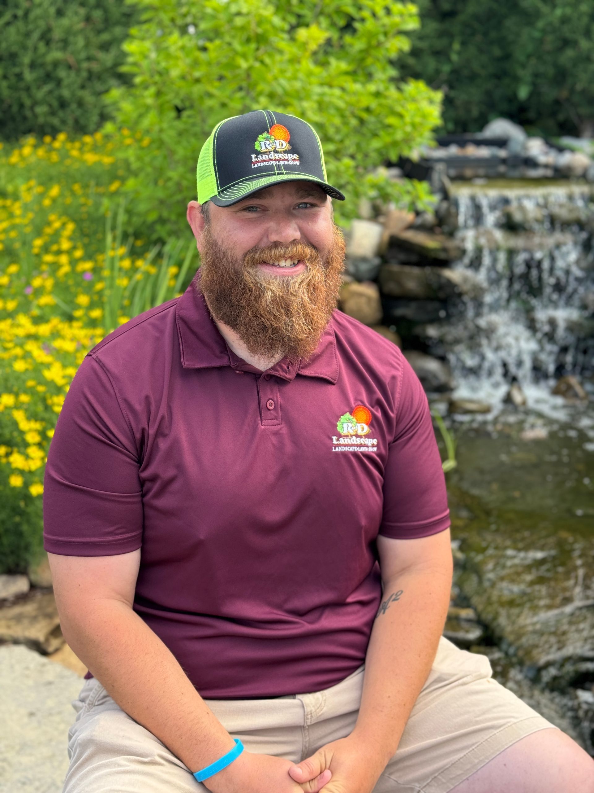A man with a beard and hat is sitting in front of a waterfall.