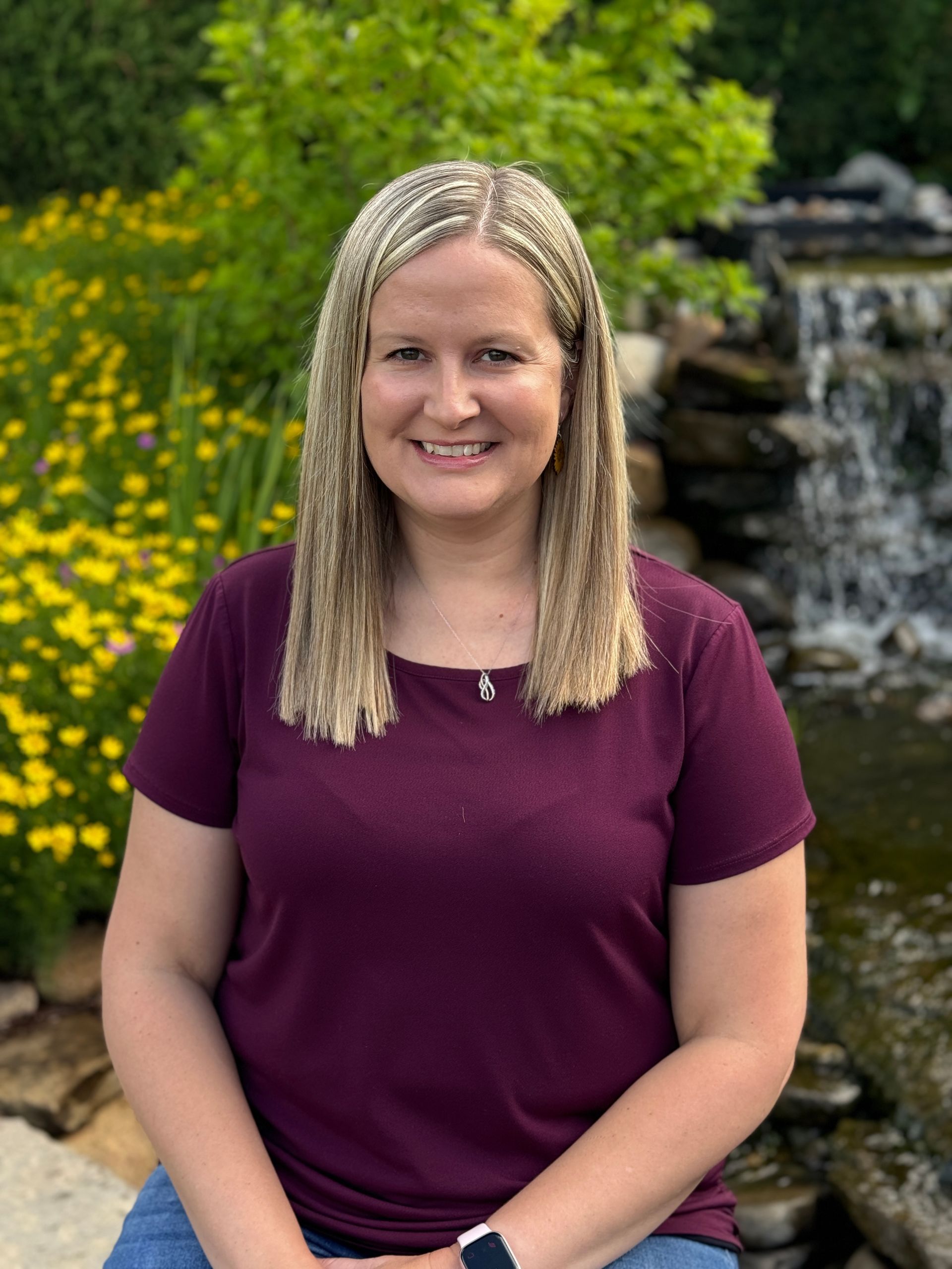A woman in a purple shirt is sitting in front of a waterfall.