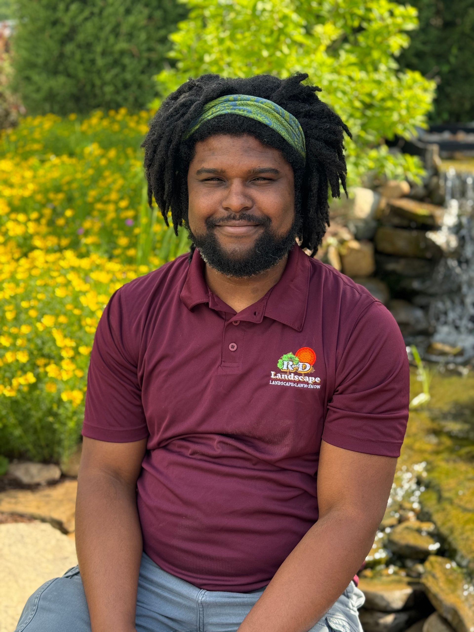 A man with dreadlocks and a beard is sitting in front of a waterfall.