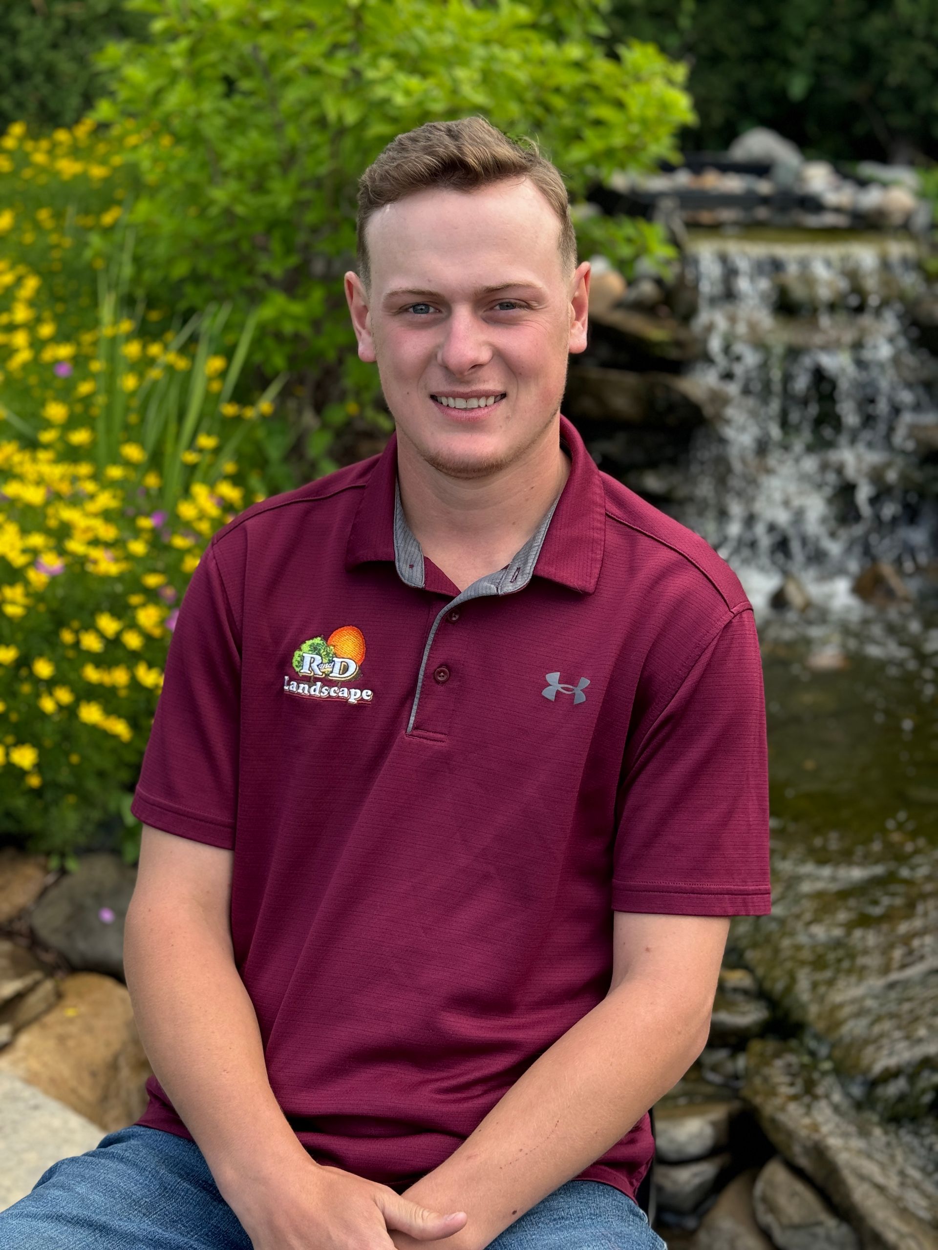 A young man in a maroon shirt is sitting in front of a waterfall.