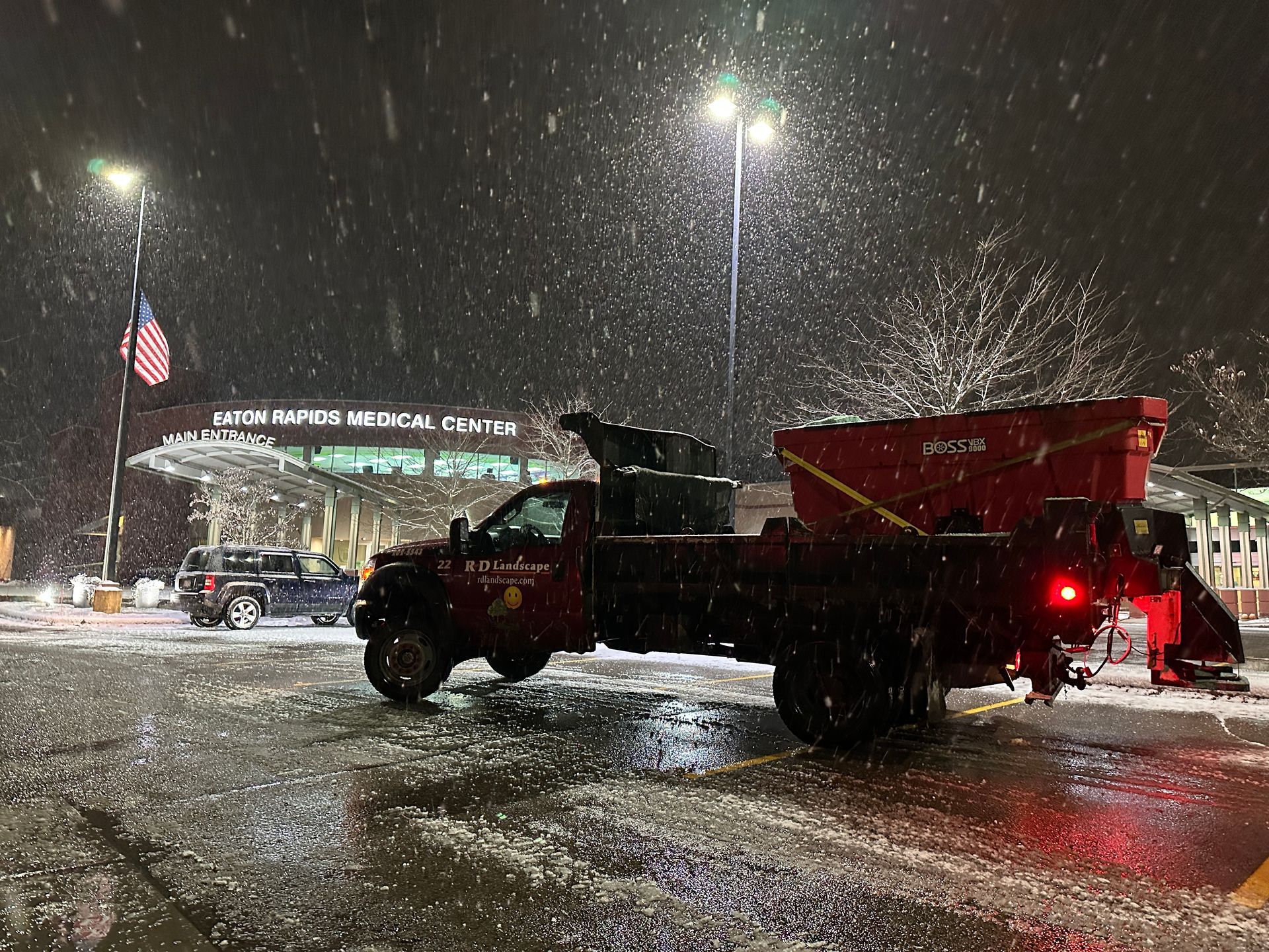 Salt truck spreading sand on a snowy road at night near a lit entrance sign