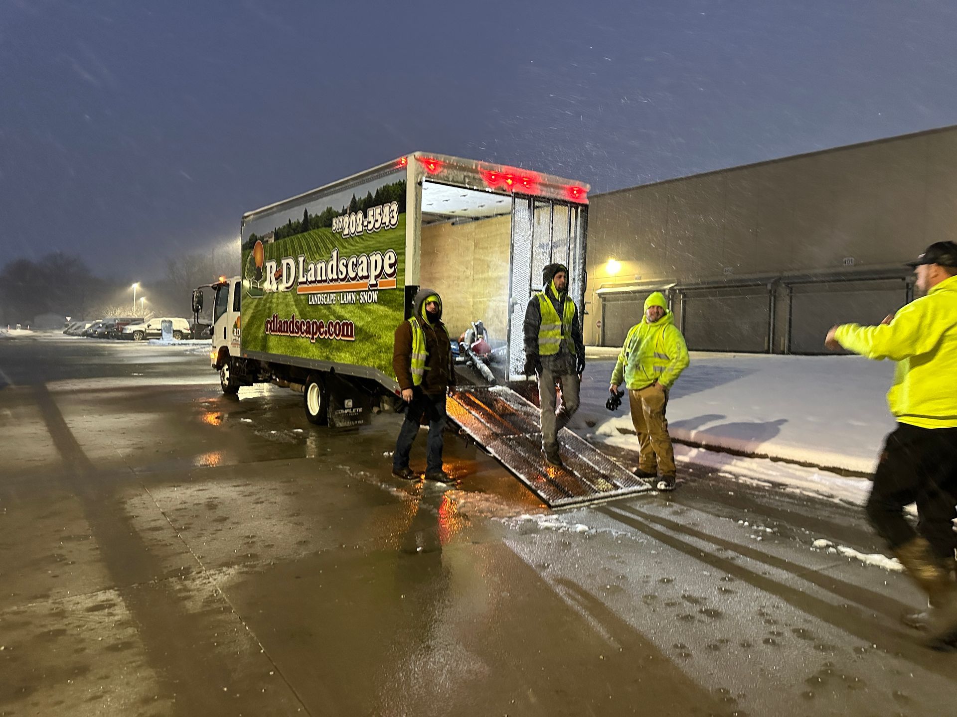 Workers in reflective gear load a large trailer in snowy parking lot at night