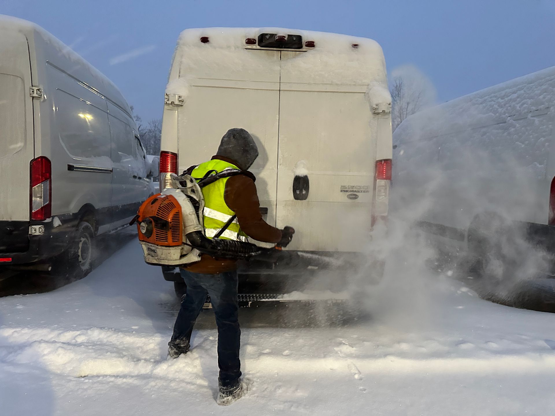 Worker in reflective vest uses a backpack blower to clear snow behind white vans.