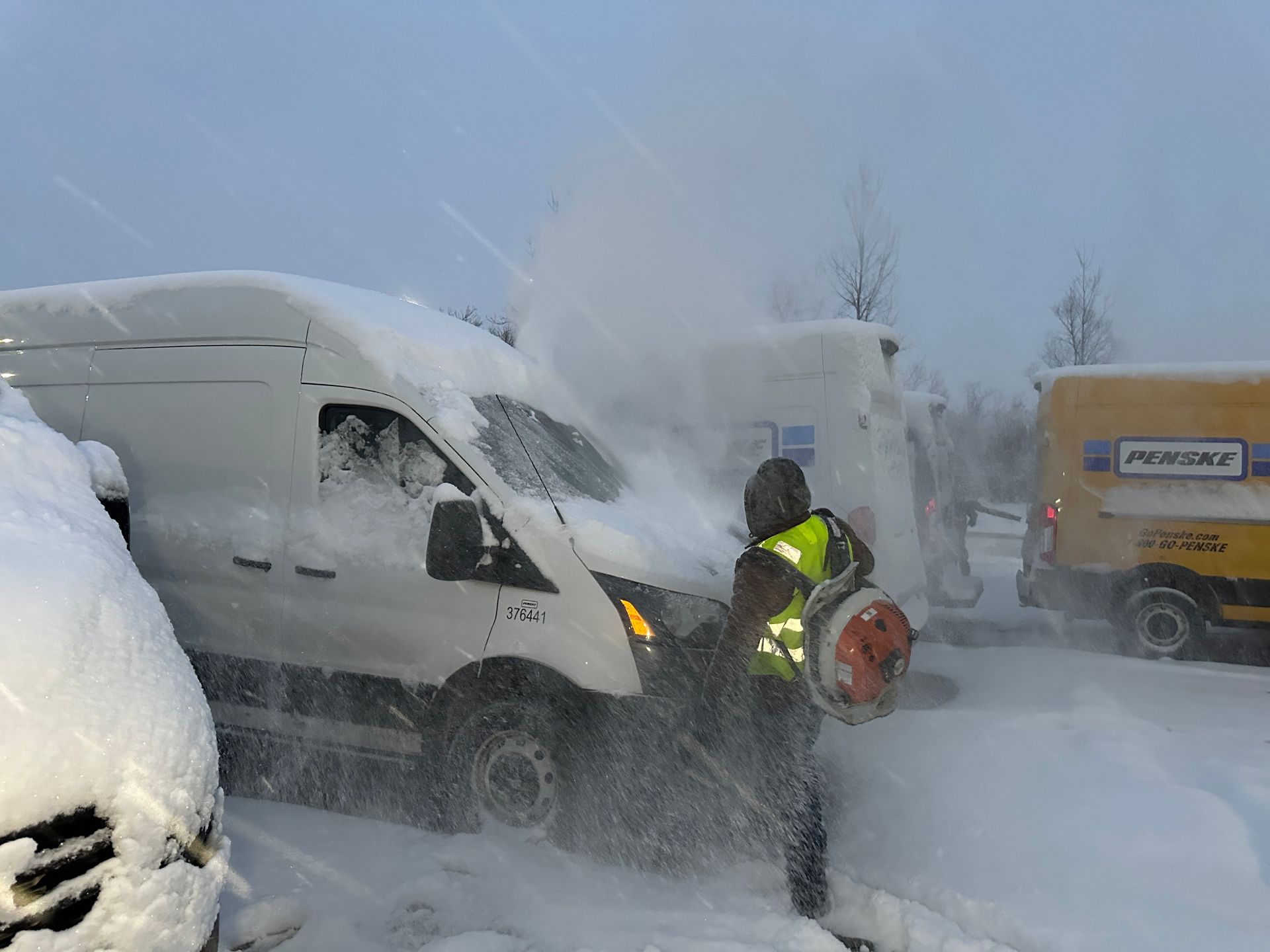 Worker in a high-visibility jacket clearing snow from a white van in a heavy snowstorm.