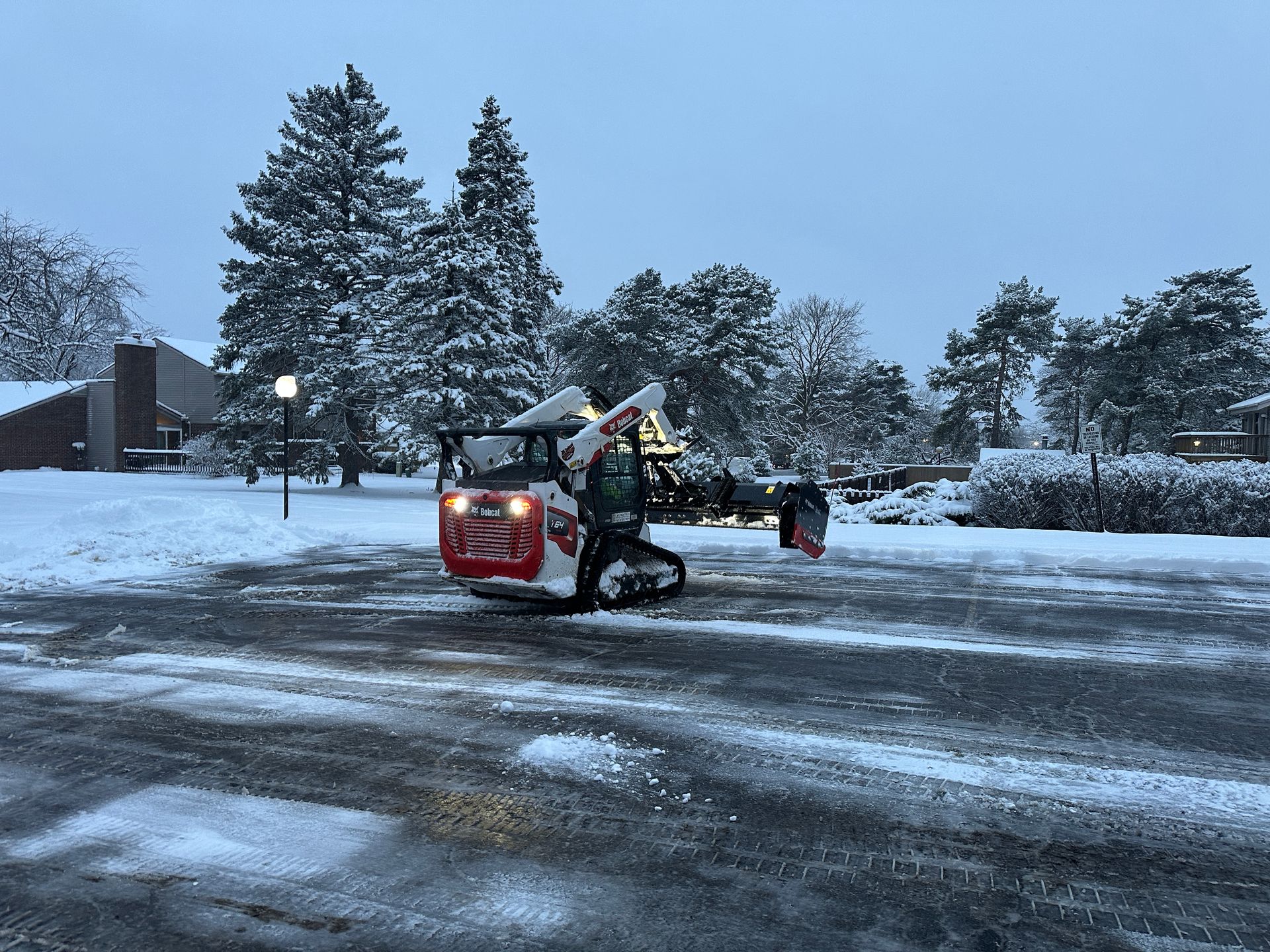 Snowplow clearing a snowy parking lot at dusk, with lit apartment buildings and snow-covered trees.