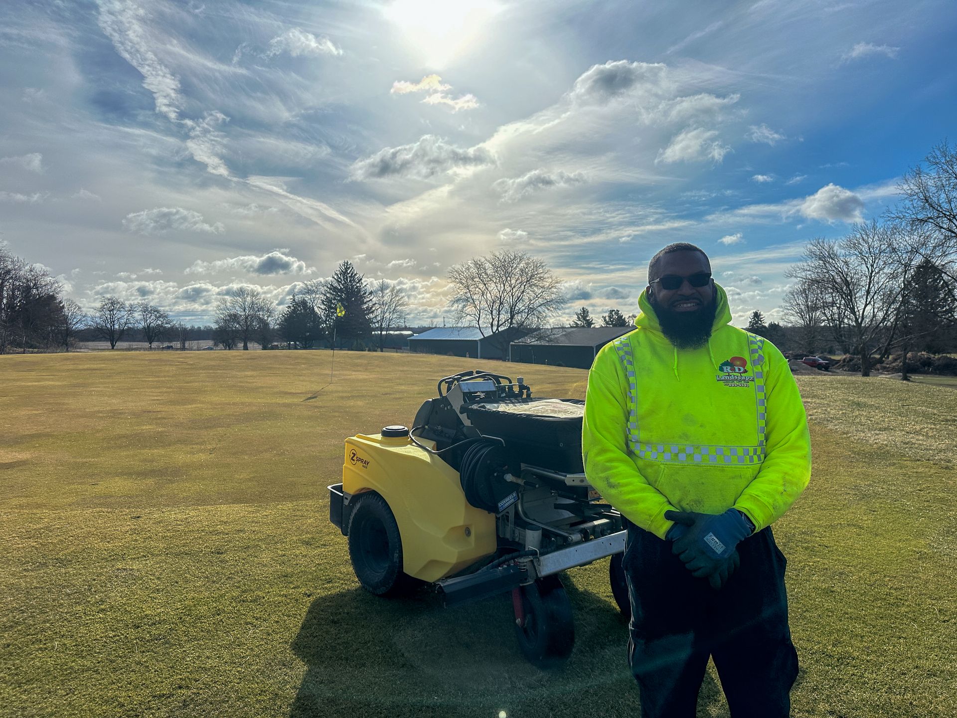 Person in a neon yellow jacket standing beside a yellow lawn machine in a grassy field under a cloudy sky