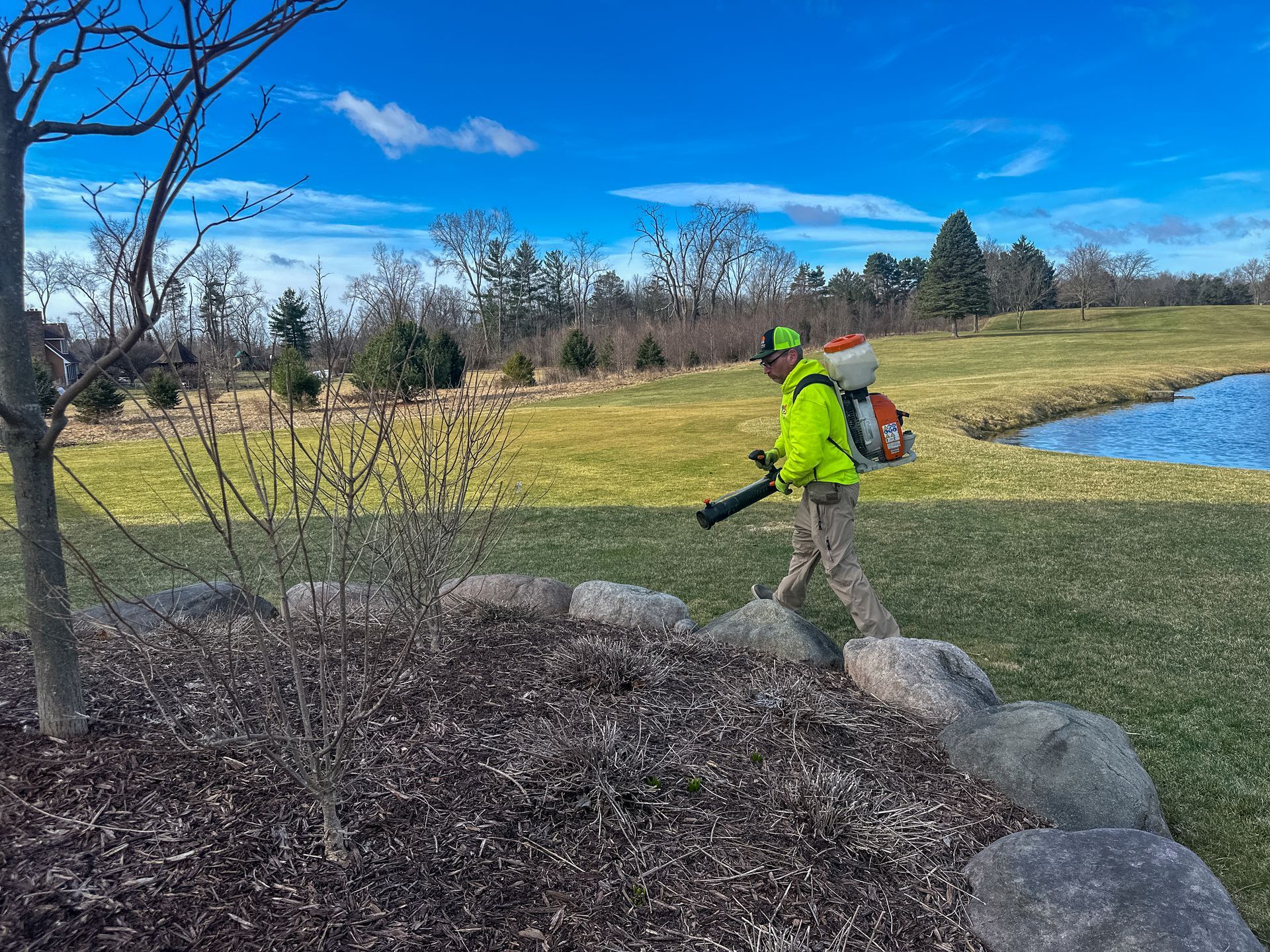 Worker in neon jacket using a leaf blower beside rocks near a pond on a grassy park path