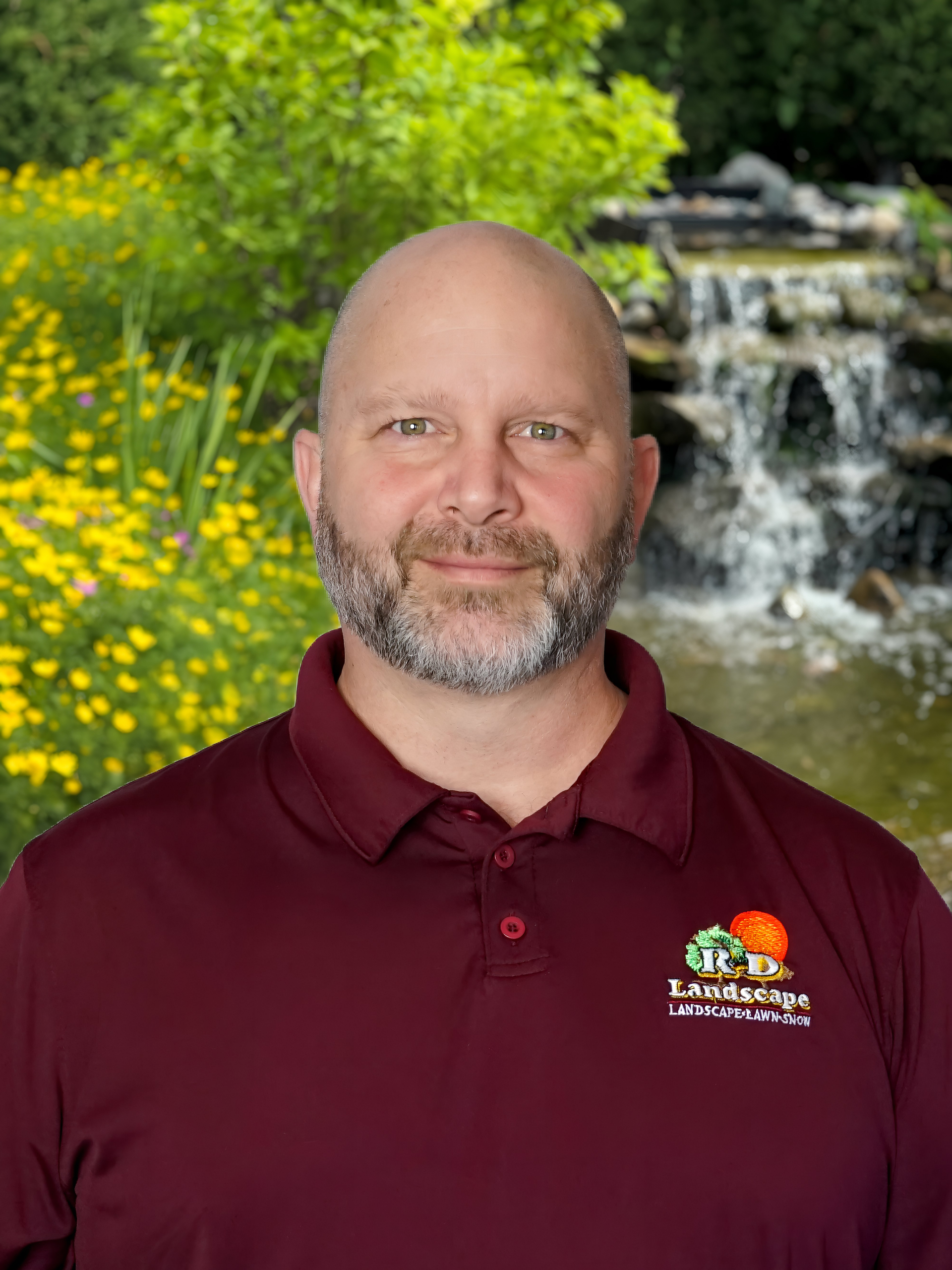 Smiling man in a maroon polo shirt with a waterfall and yellow flowers in the background