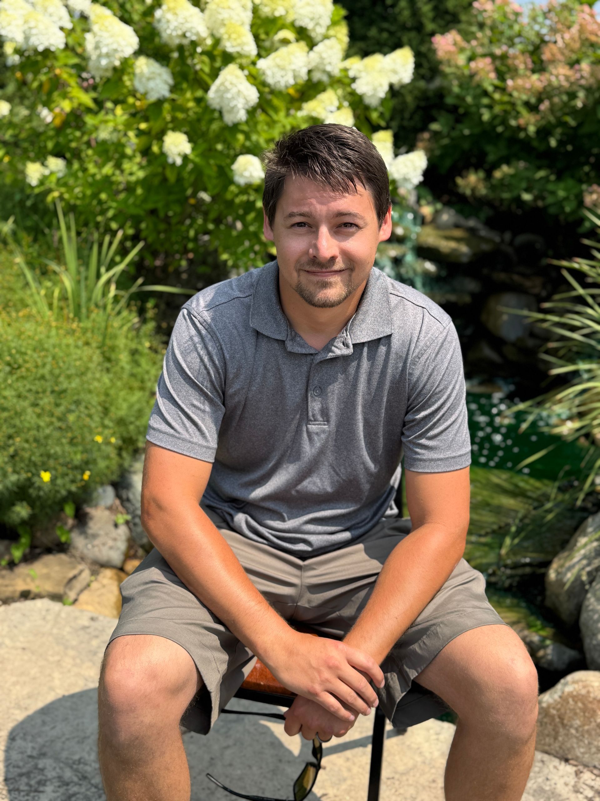 Man sitting on a rock in a garden, smiling in a gray polo and shorts.