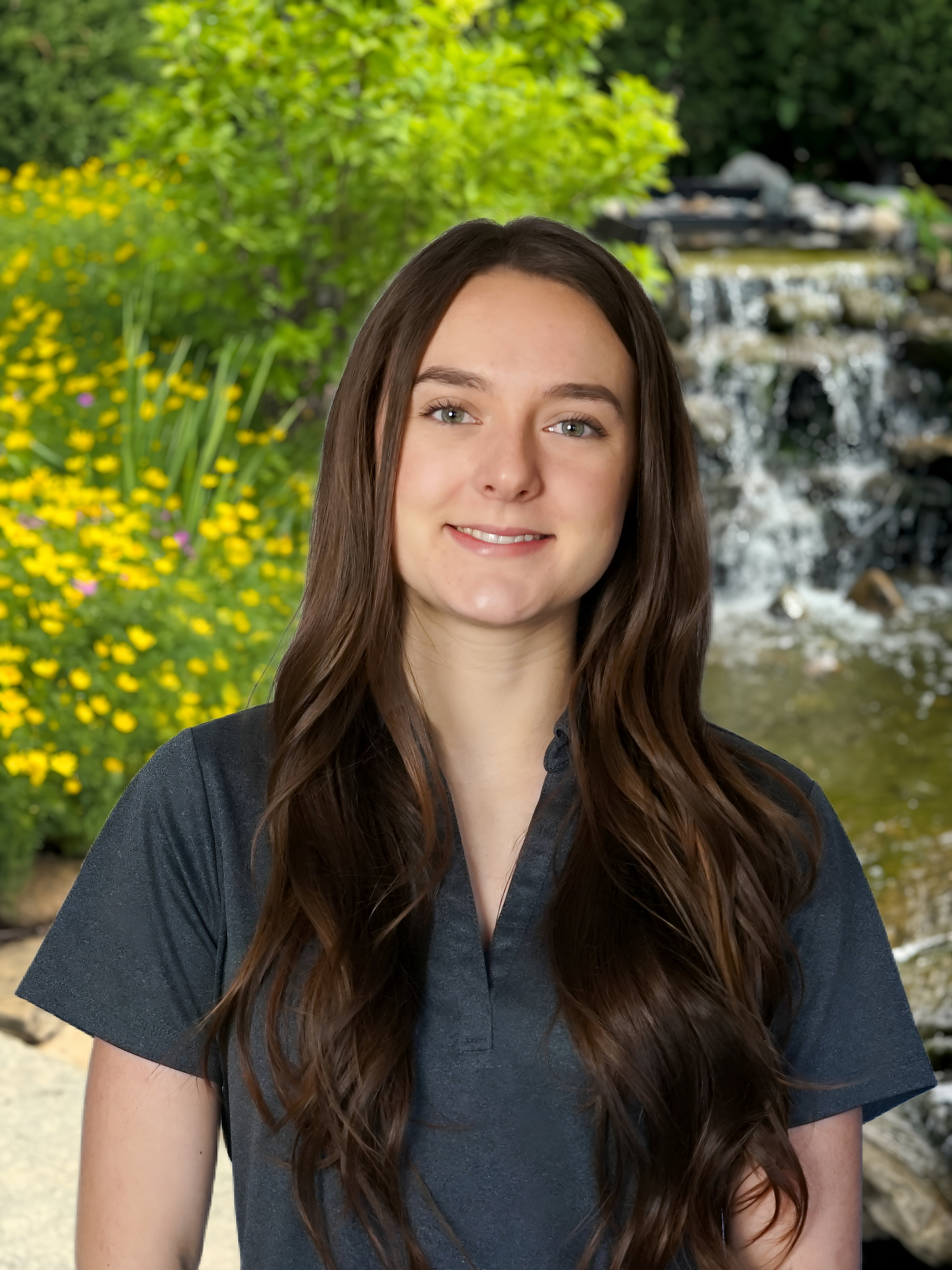 Portrait of a smiling woman with long dark hair in a garden beside yellow flowers and a waterfall