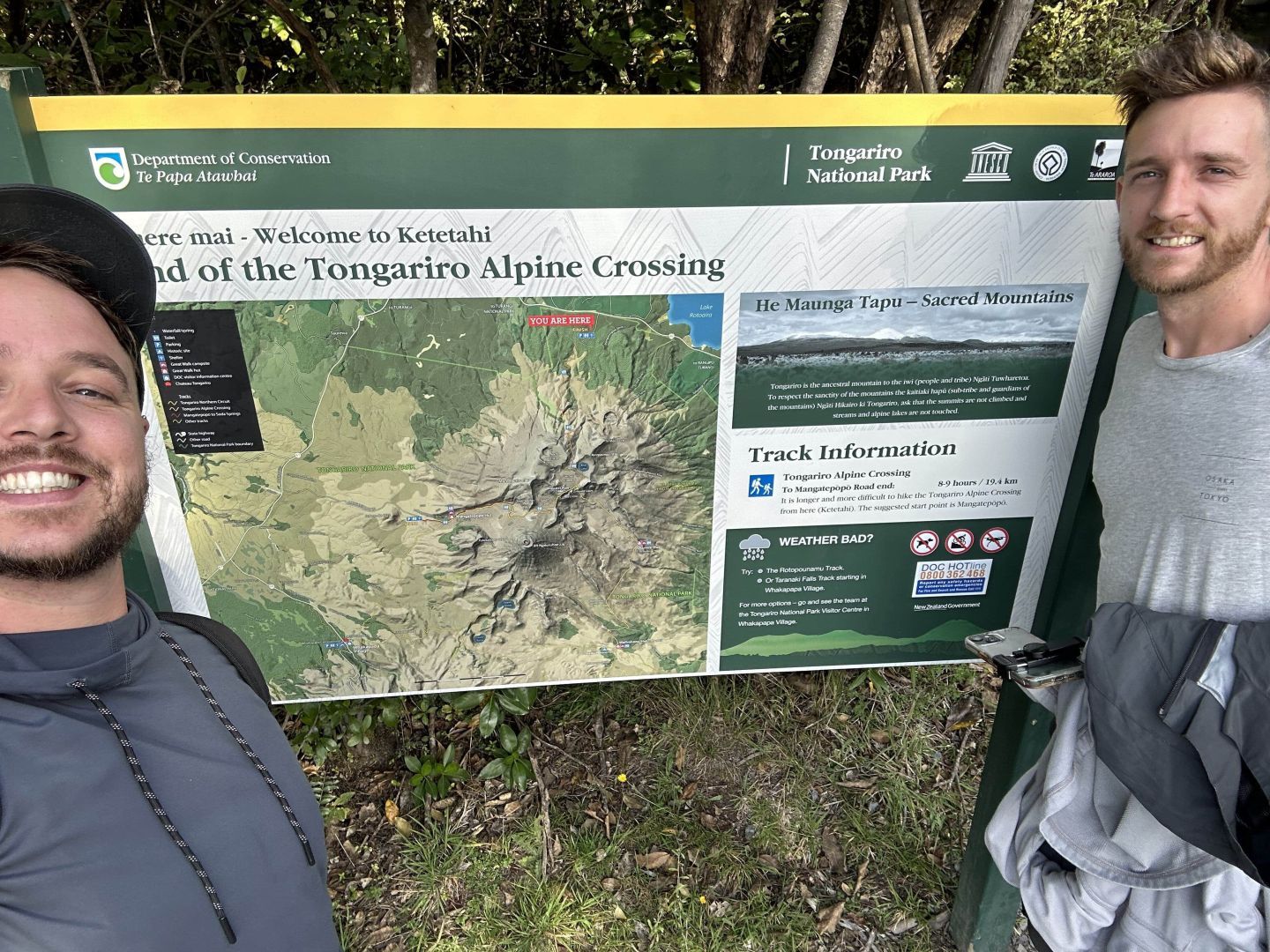 Two people taking a selfie on front of the Tongariro Alpine Crossing sign at the start of the track