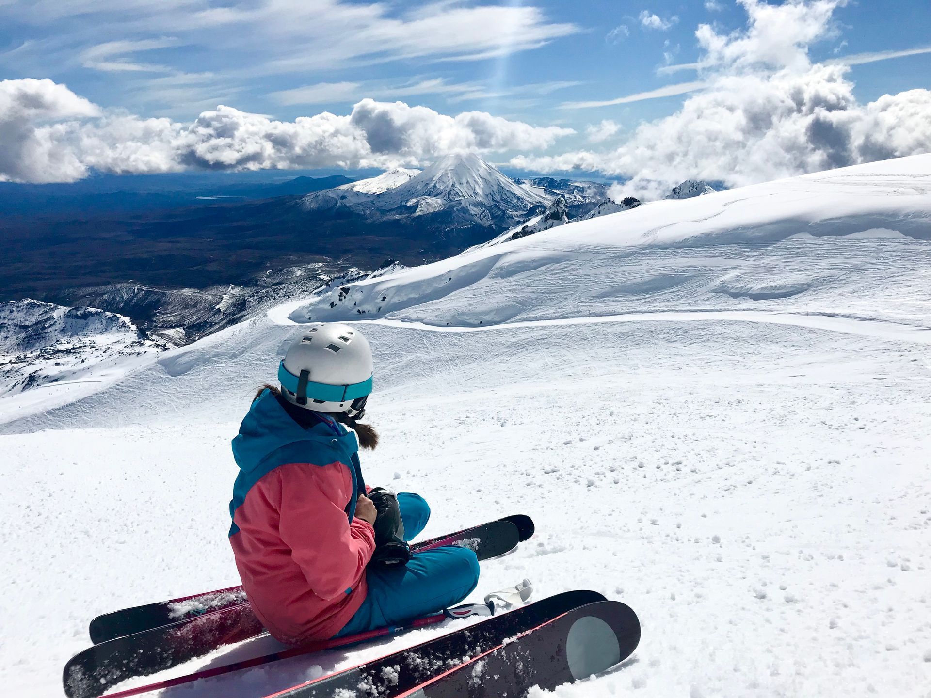 Skier in pink jacket sitting on snowy slope overlooking mountains and clouds