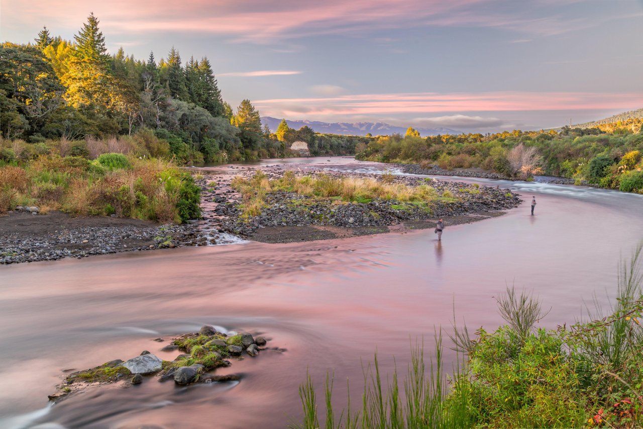 Pink sunset over a winding river with rocky banks and lush green trees.