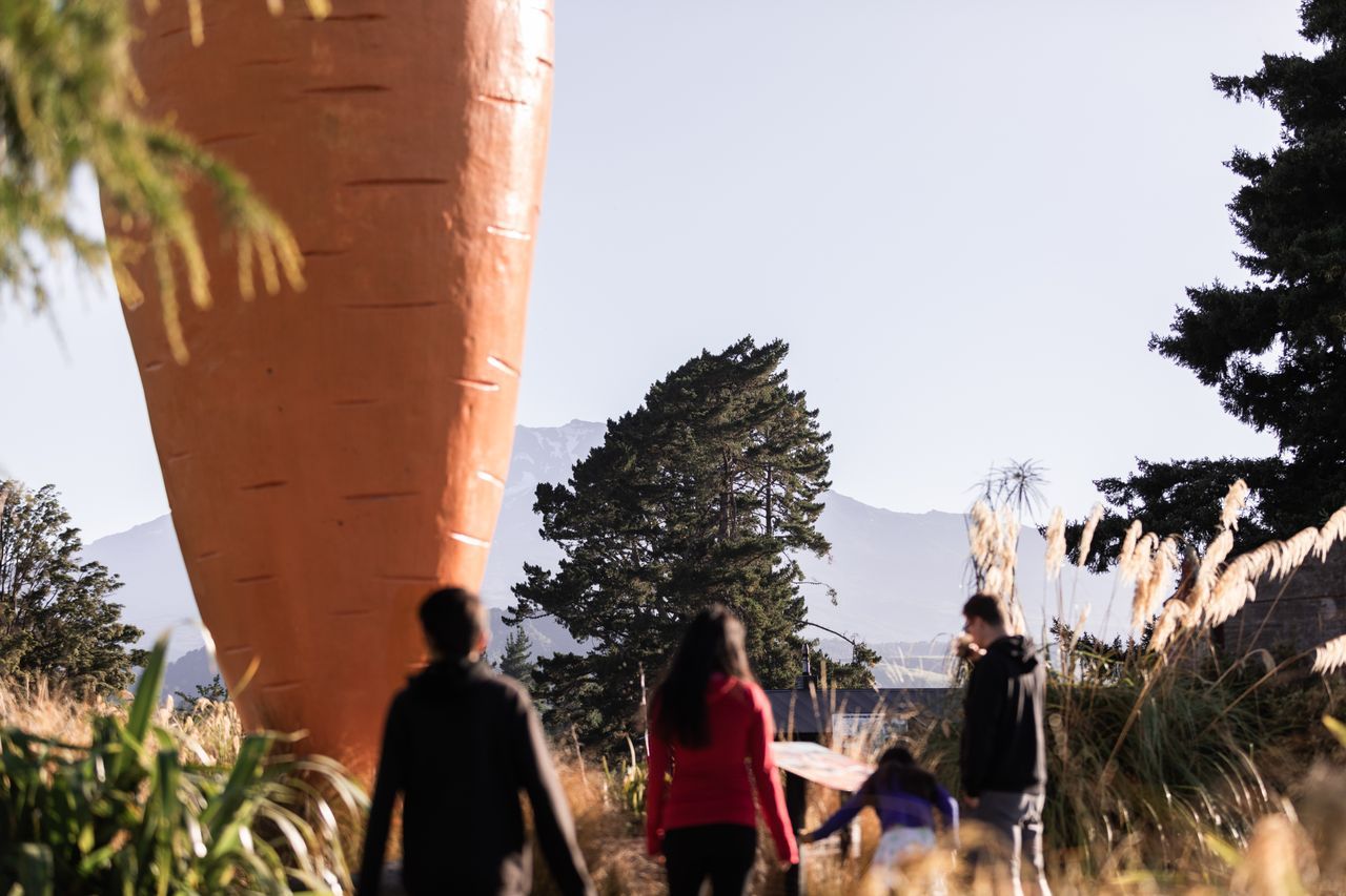 People walking near a large orange sculpture outdoors with trees and mountains in the background