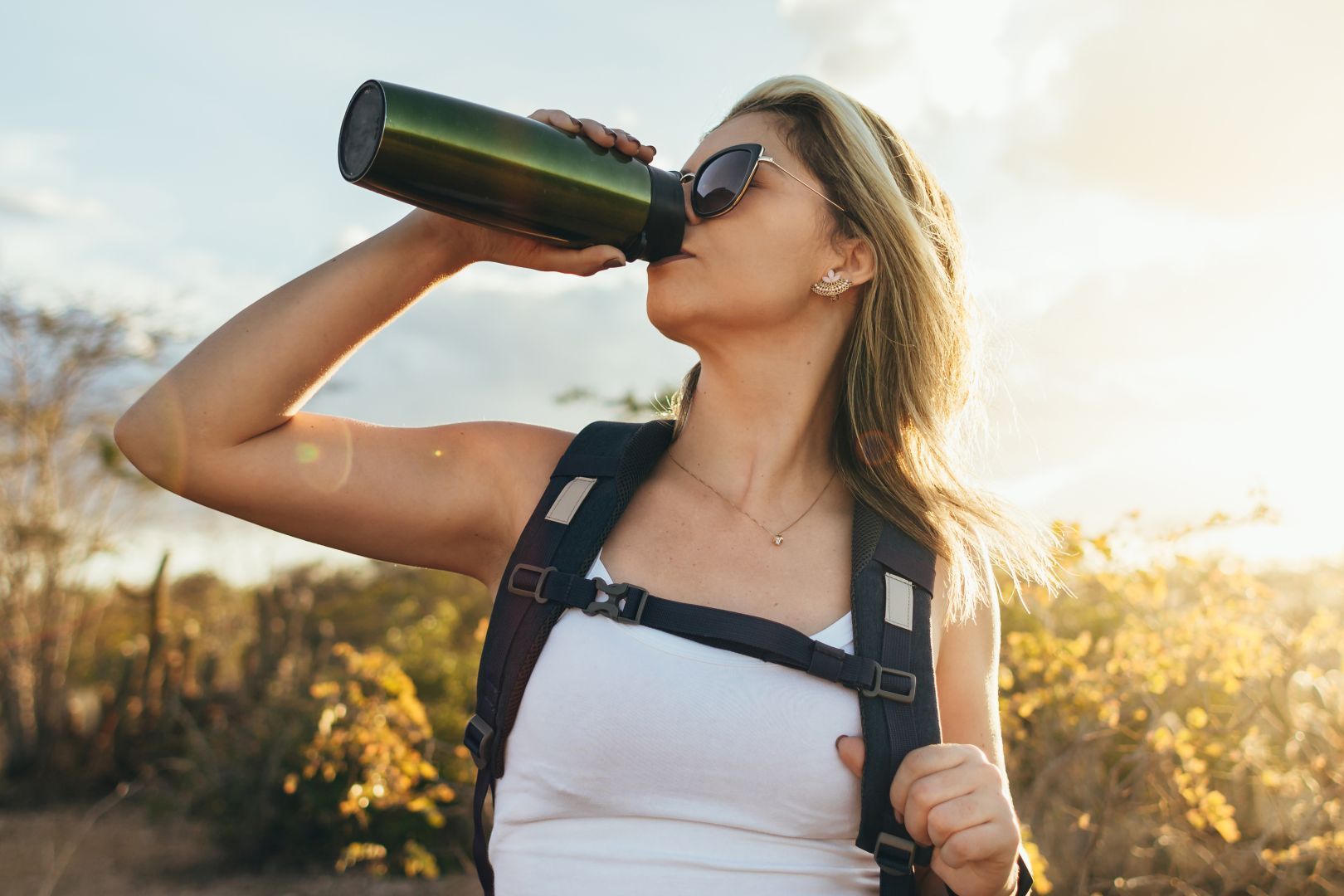 Woman in sunglasses drinking from a bottle outdoors at sunset, wearing a white top and backpack straps