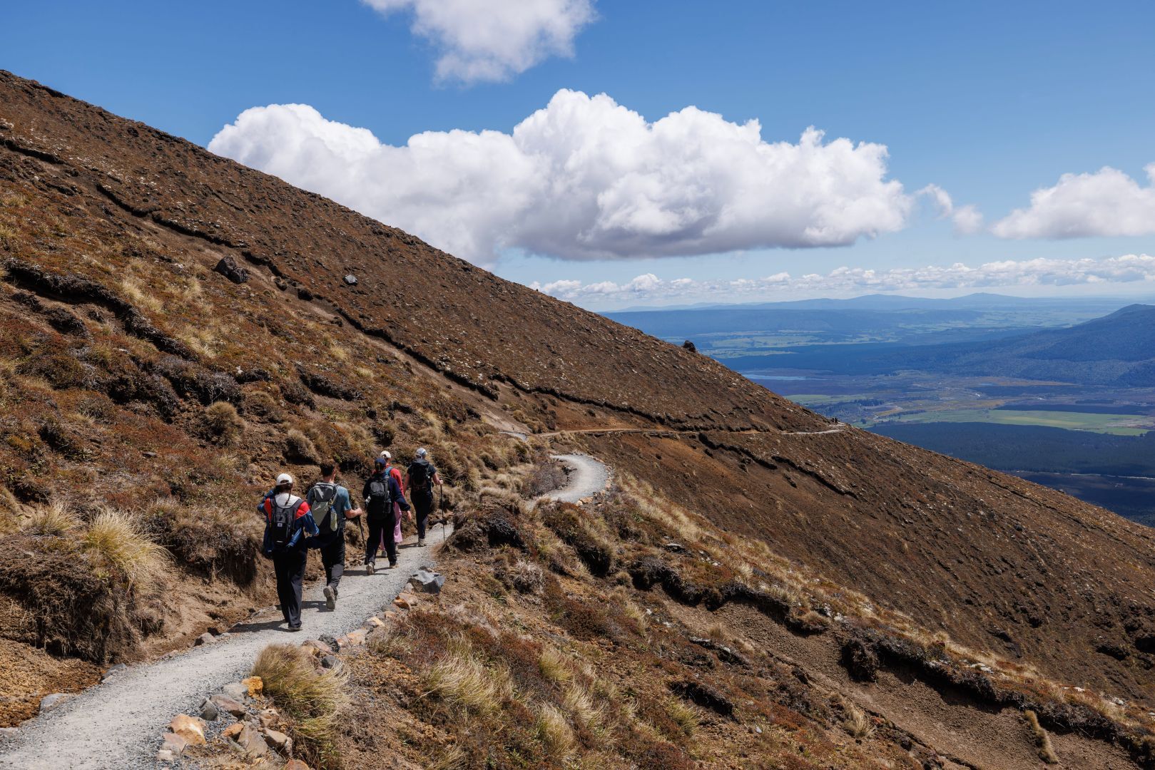 Hikers on a narrow mountain trail beside a brown hillside under a blue sky with clouds