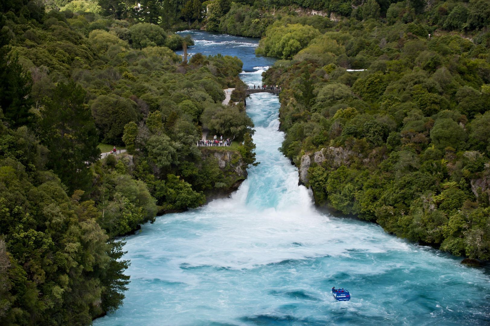 Wide turquoise waterfall rushing through dense green forest with a small blue raft in the foreground