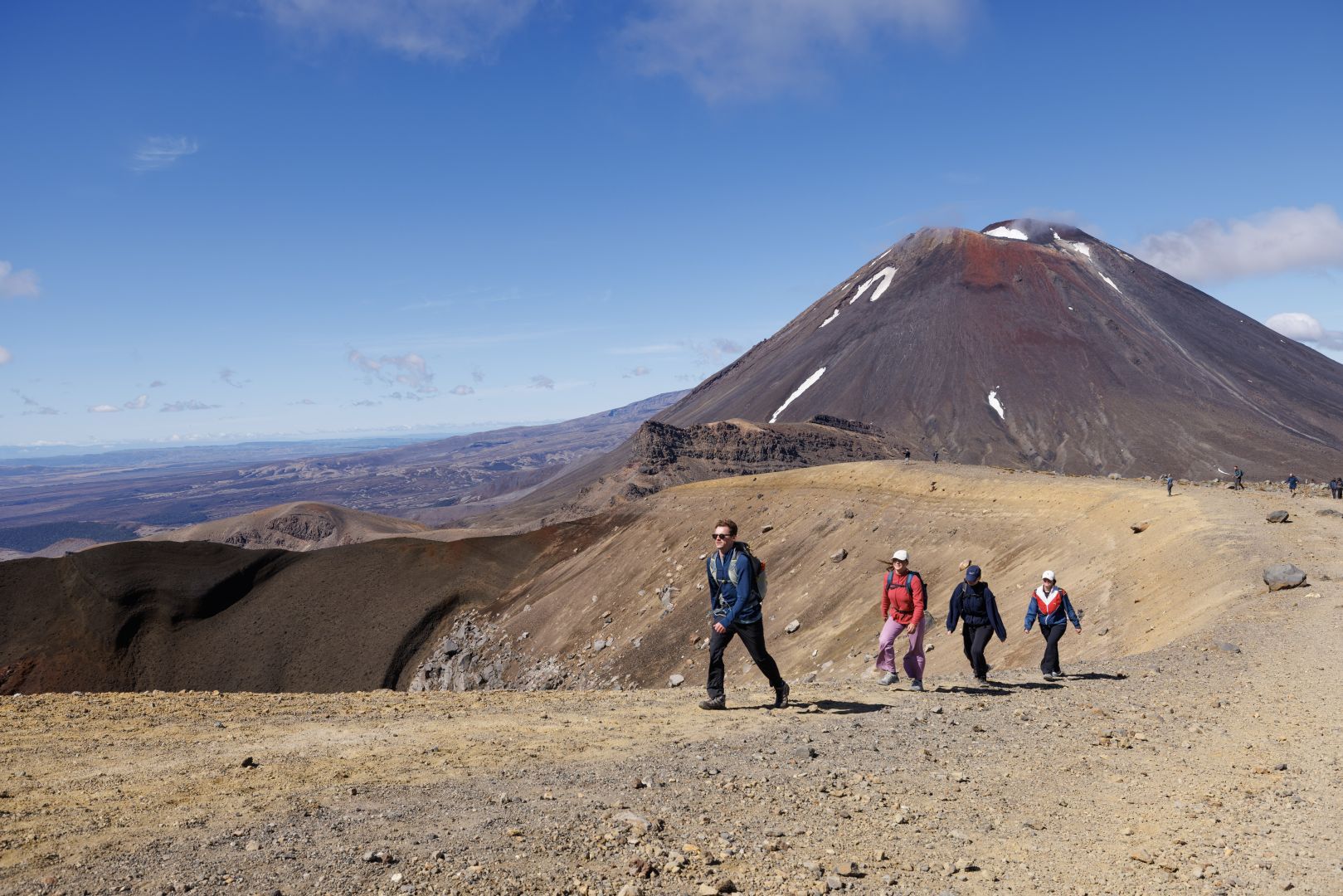 Hikers crossing a barren volcanic landscape with a large mountain under a blue sky