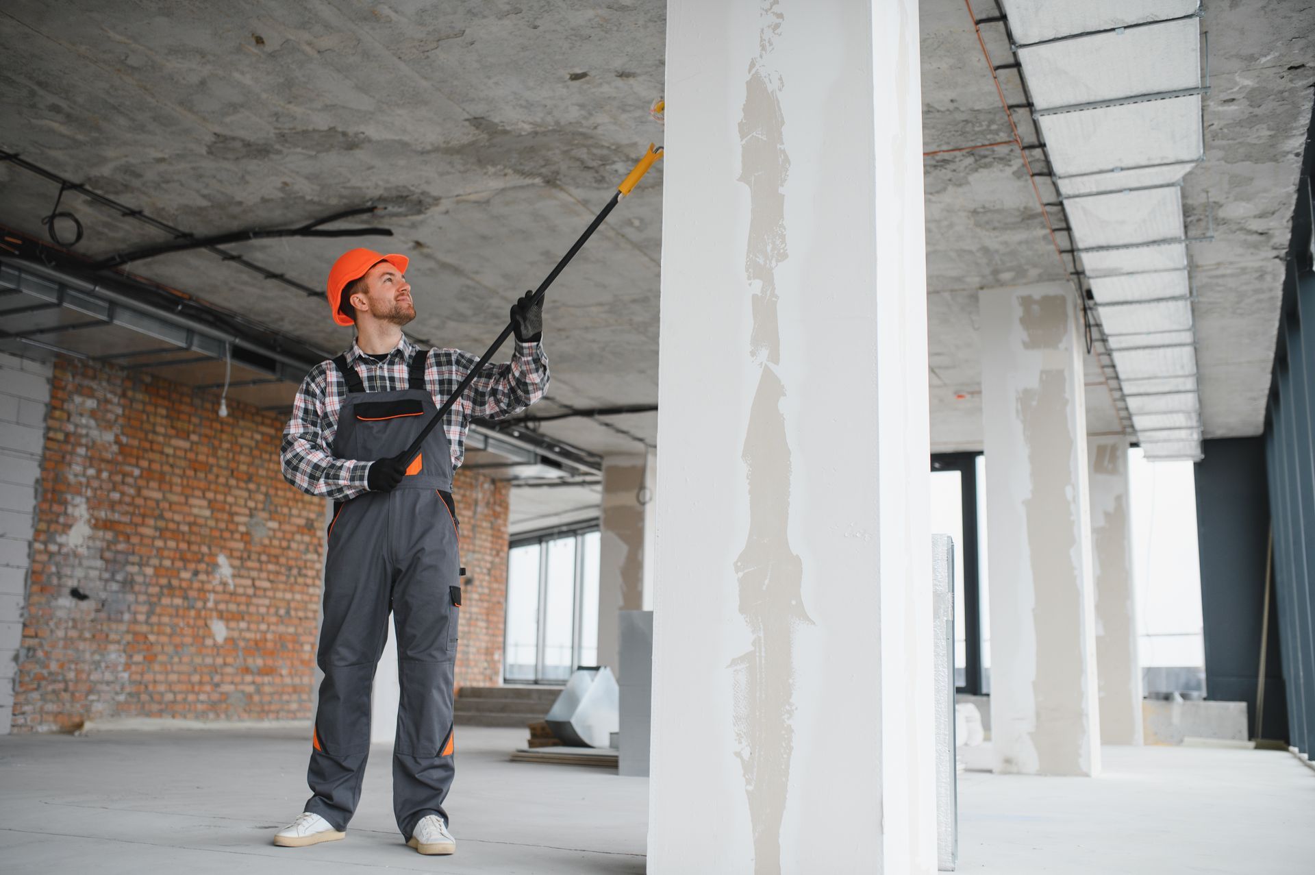Construction worker spraying a ceiling with a long pole in an unfinished building interior
