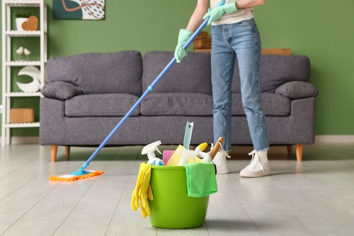 Person mopping a living room floor beside a green cleaning bucket and supplies