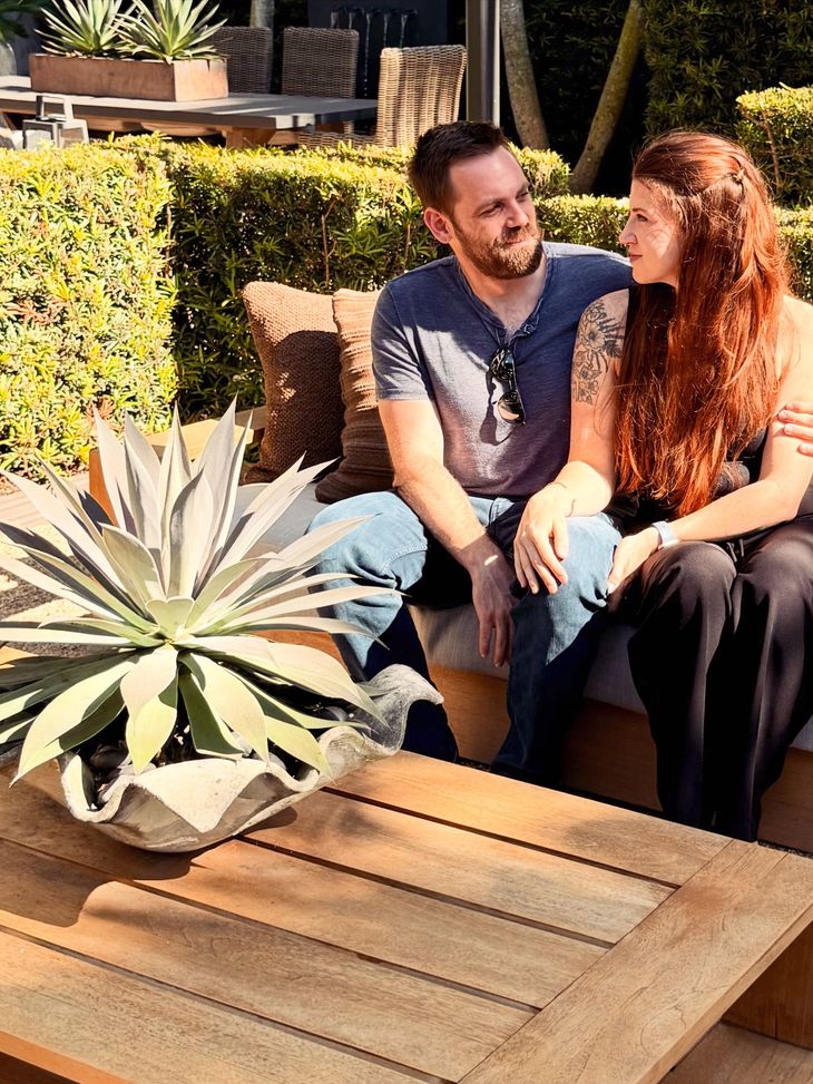Two people sit on a patio bench beside a large potted succulent, smiling in bright sunlight.
