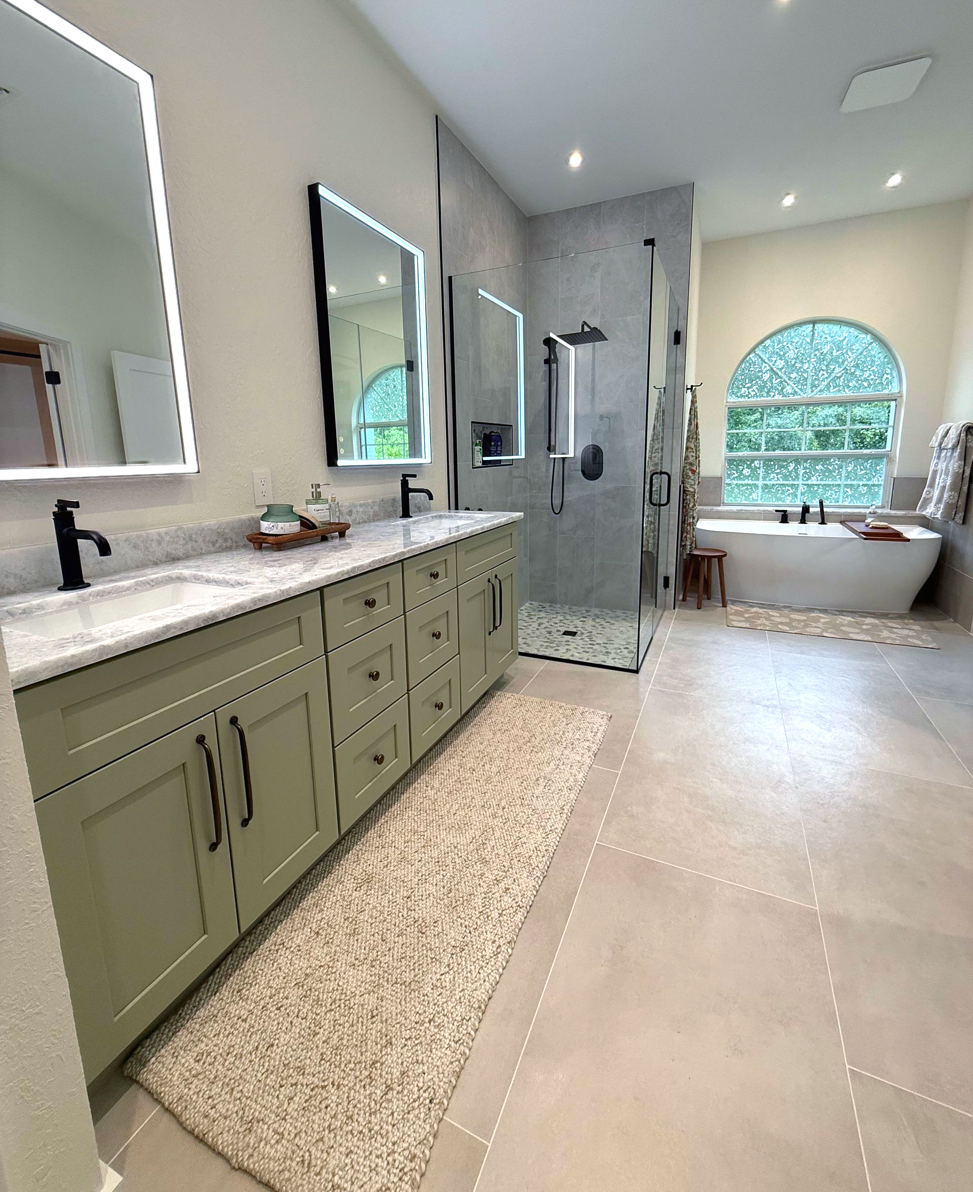 Modern bathroom with green vanity, large mirrors, walk-in shower, and freestanding tub by arched window.