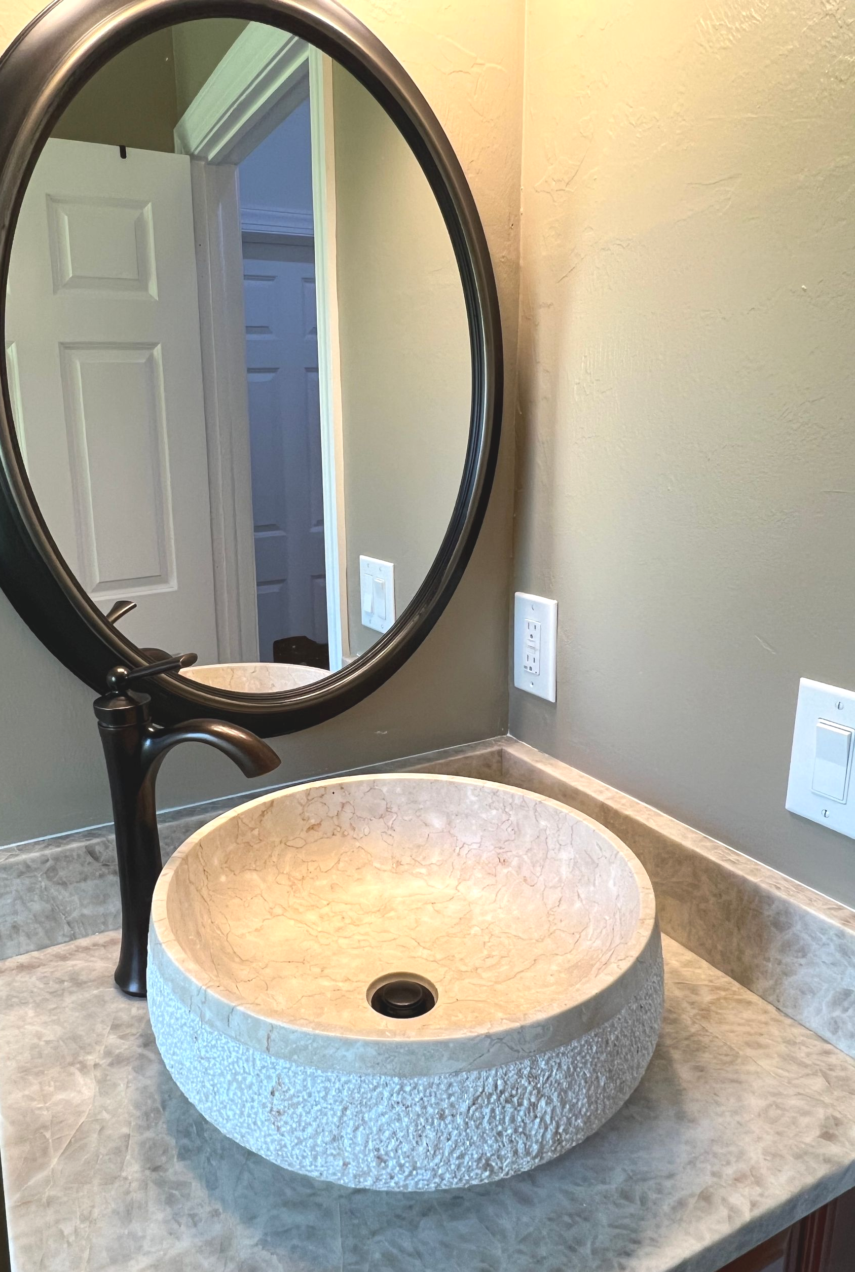 Bathroom vanity with round stone vessel sink, black faucet, and oval mirror on a gray countertop.