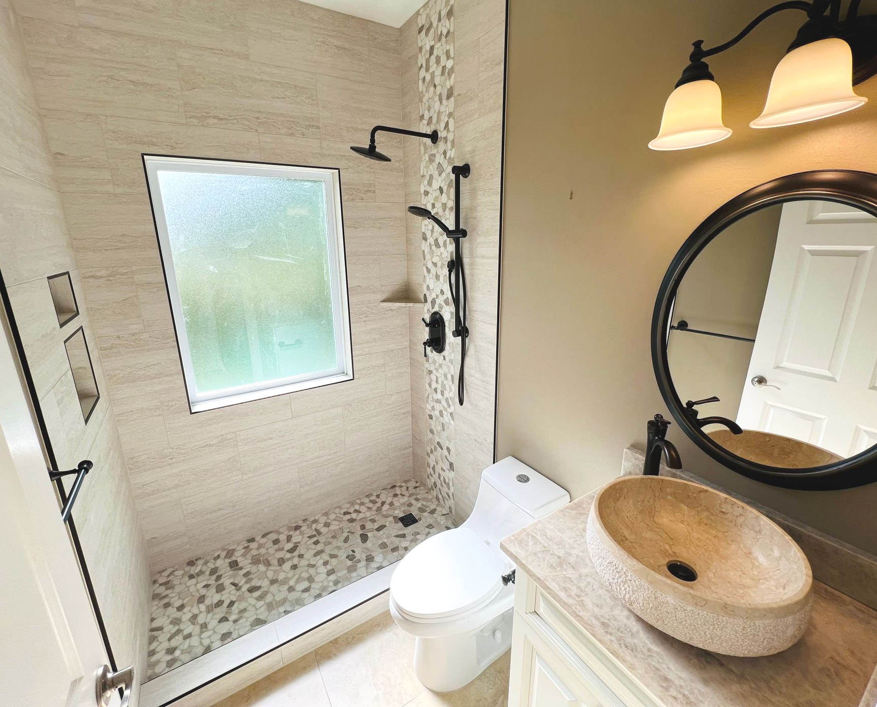 Bright bathroom with tiled shower, frosted window, sink, toilet, and round mirror.