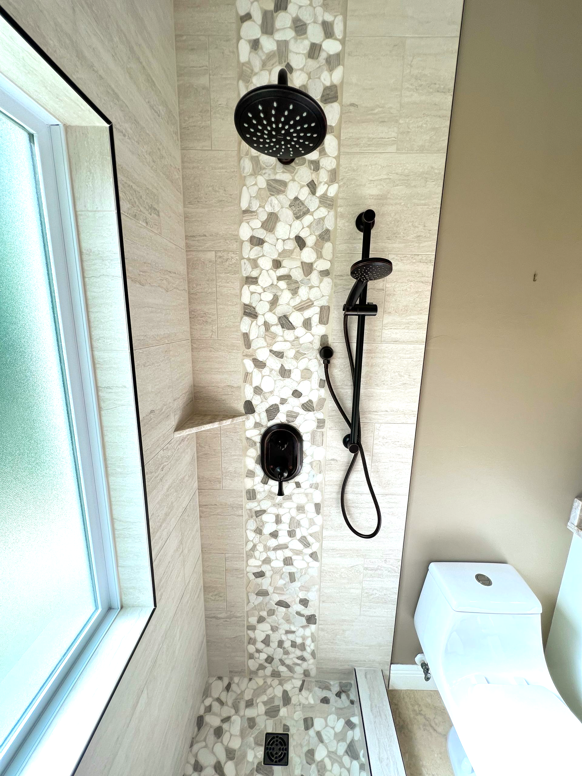 Bathroom shower with mosaic tile, black showerhead and hose, next to a toilet and window.