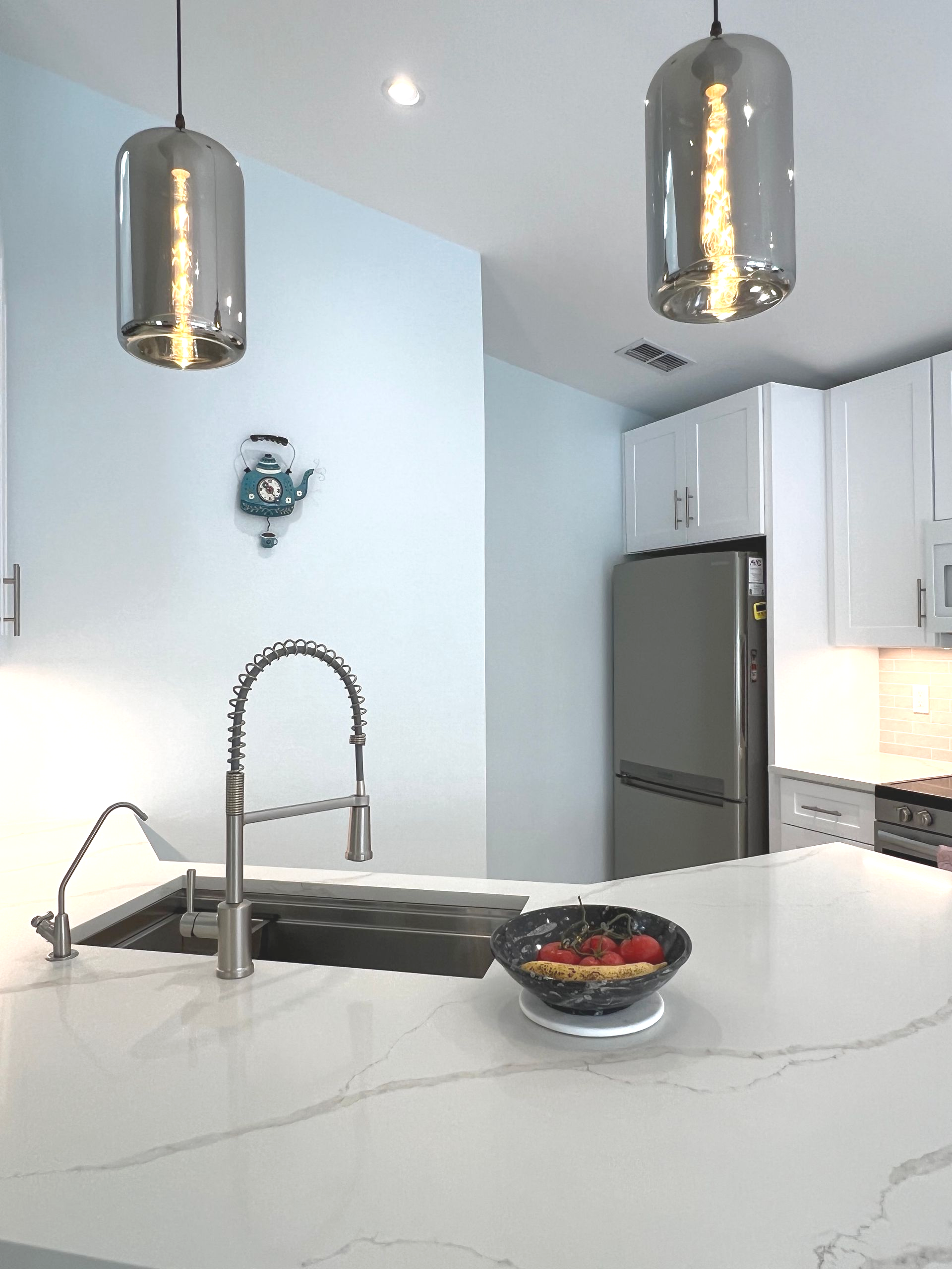 Modern white kitchen with marble island, stainless sink, pendant lights, and bowl of fruit