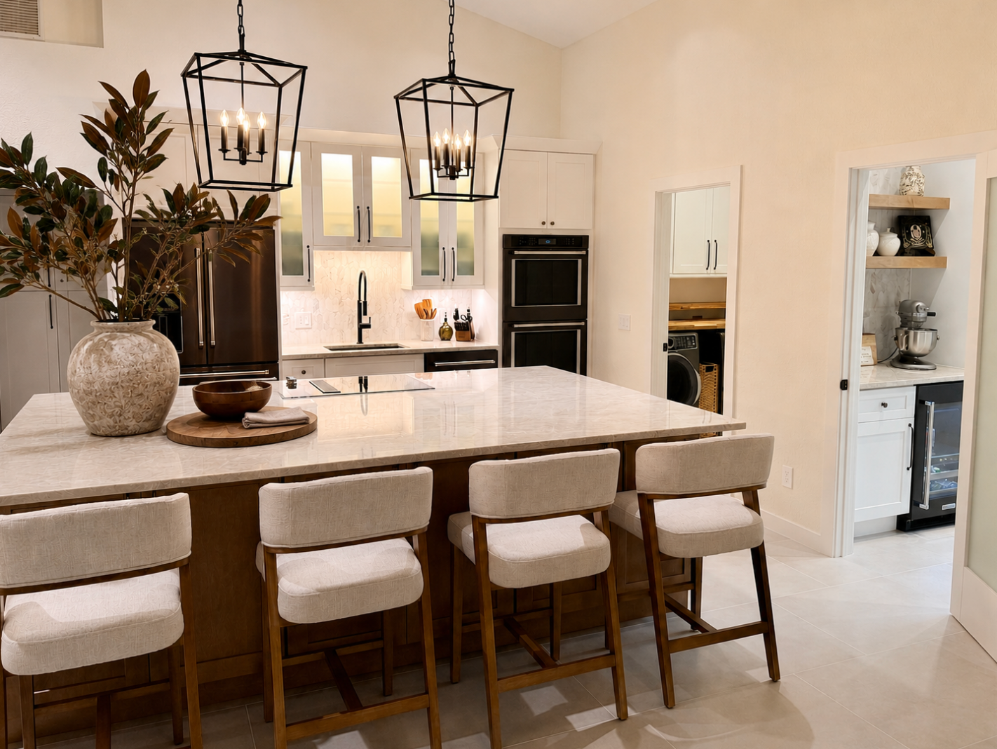 Modern white kitchen with island, cushioned barstools, pendant lights, and built-in ovens