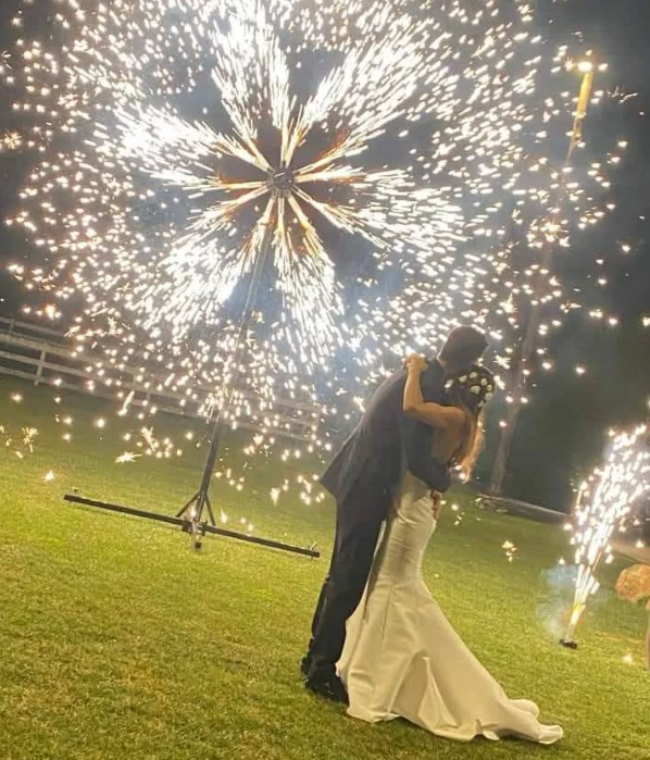 Gli sposi si abbracciano mentre i fuochi d'artificio esplodono sopra di loro durante una cerimonia nuziale notturna su un prato erboso.
