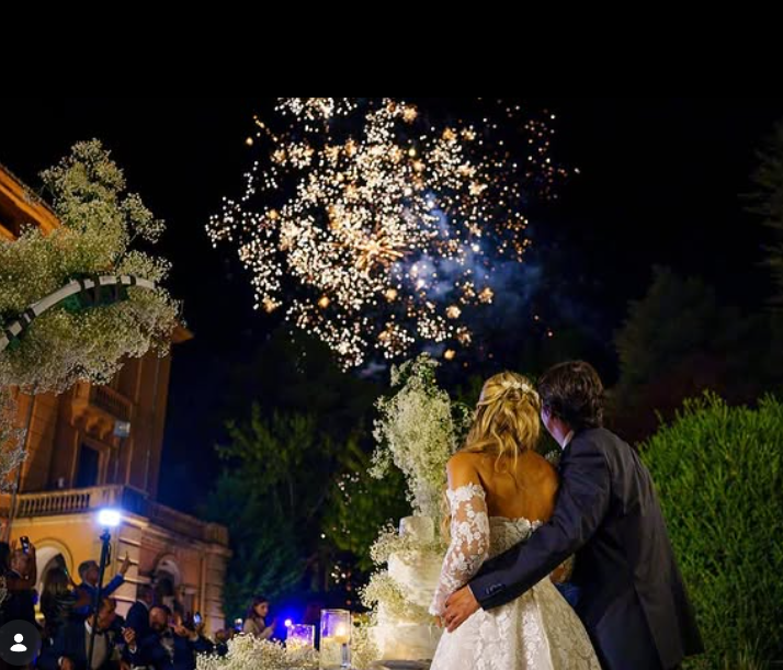 Gli sposi guardano i fuochi d'artificio. Sono in piedi vicino a una torta a più piani, con un edificio decorato e fogliame sullo sfondo.