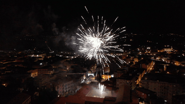 I fuochi d'artificio esplodono nel cielo notturno sopra una città, illuminando gli edifici con una luce bianca e dorata.
