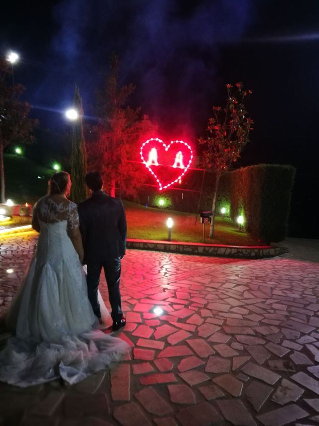 Una coppia, di spalle alla telecamera, sta in piedi davanti a un grande cuore illuminato con le iniziali, in un giardino notturno.