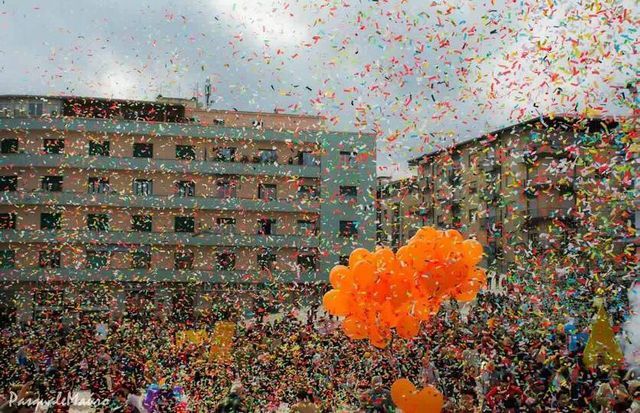 Festa di strada piena di coriandoli; palloncini arancioni fluttuano sopra la folla densa e gli edifici.